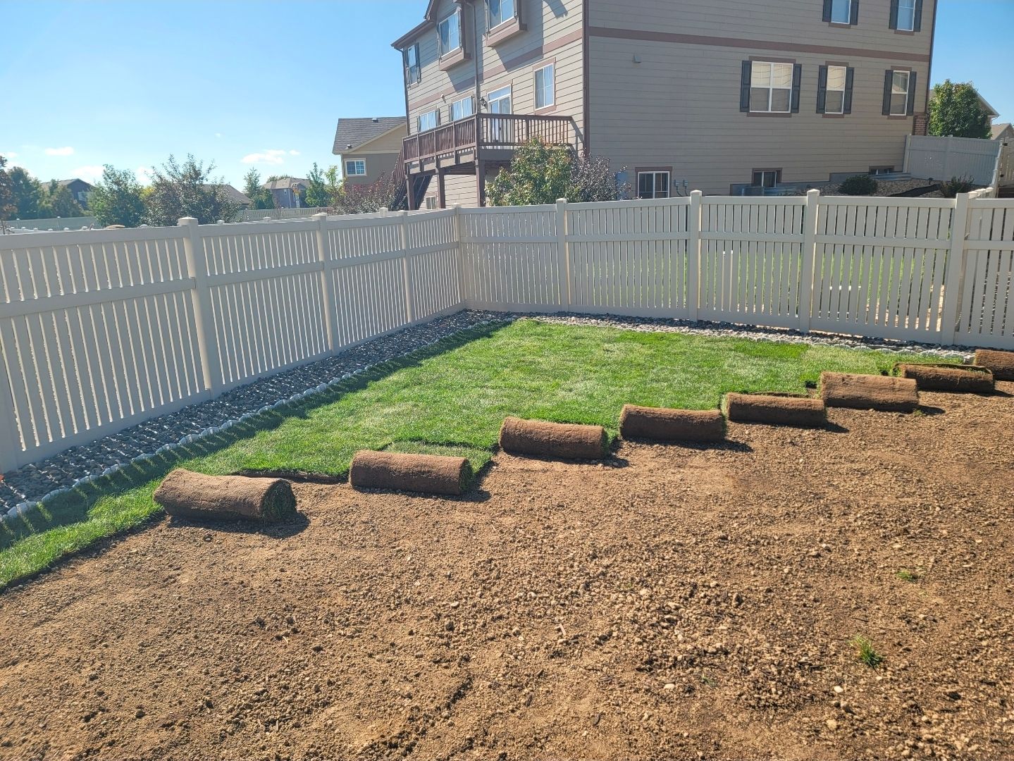 A partially sodded backyard featuring a white privacy fence and a two-story house in the background.