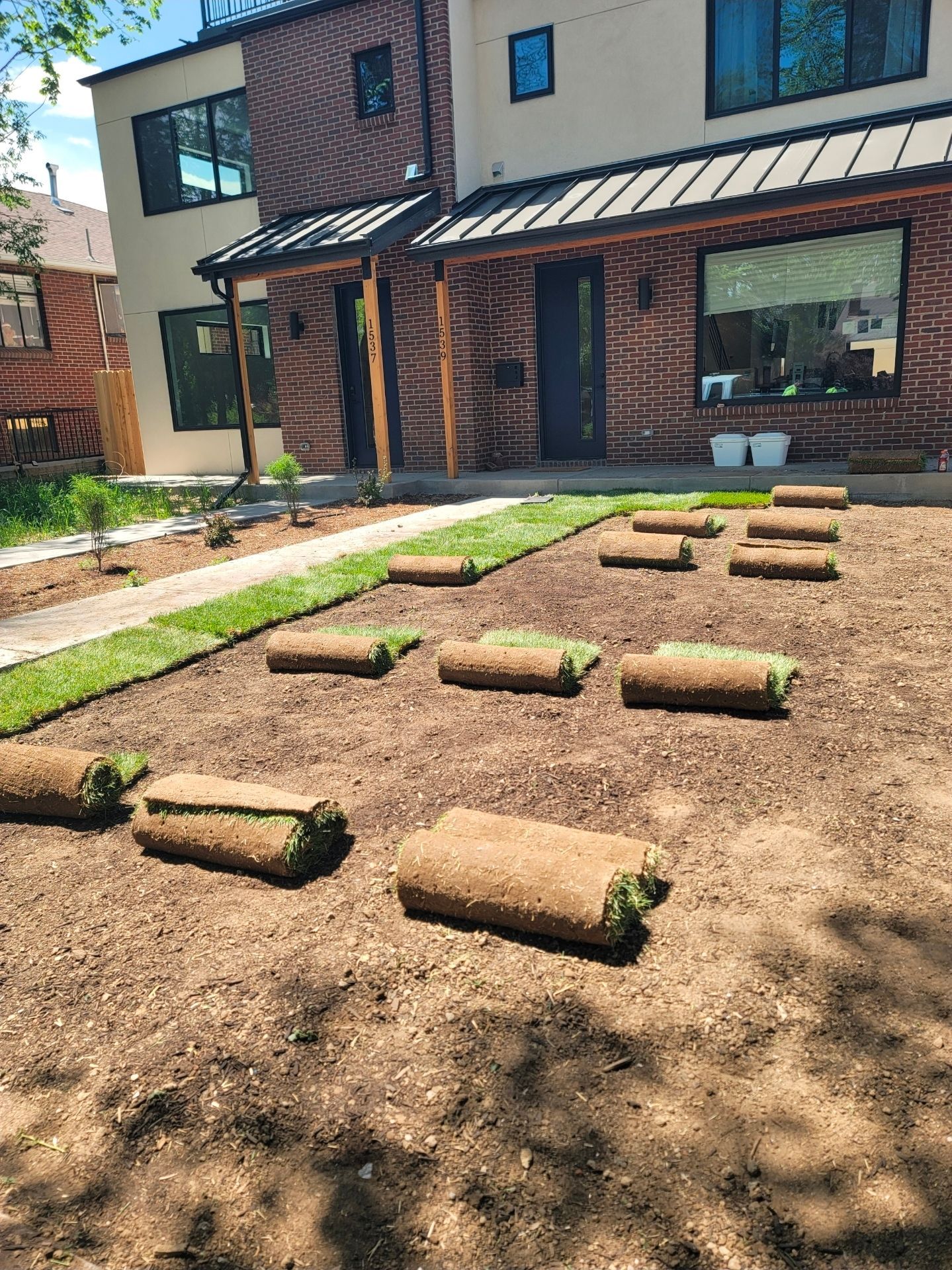 Rolls of sod spaced across a dirt yard in front of a modern two-story home with a brick and beige exterior.