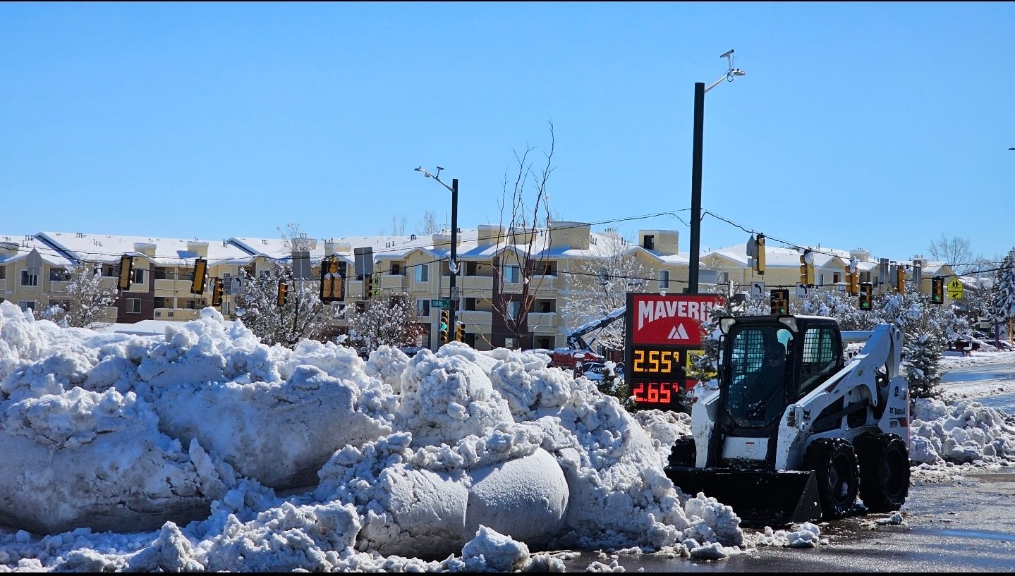 A white skid-steer loader clears large piles of snow in a parking lot next to a gas station sign under a clear blue sky.