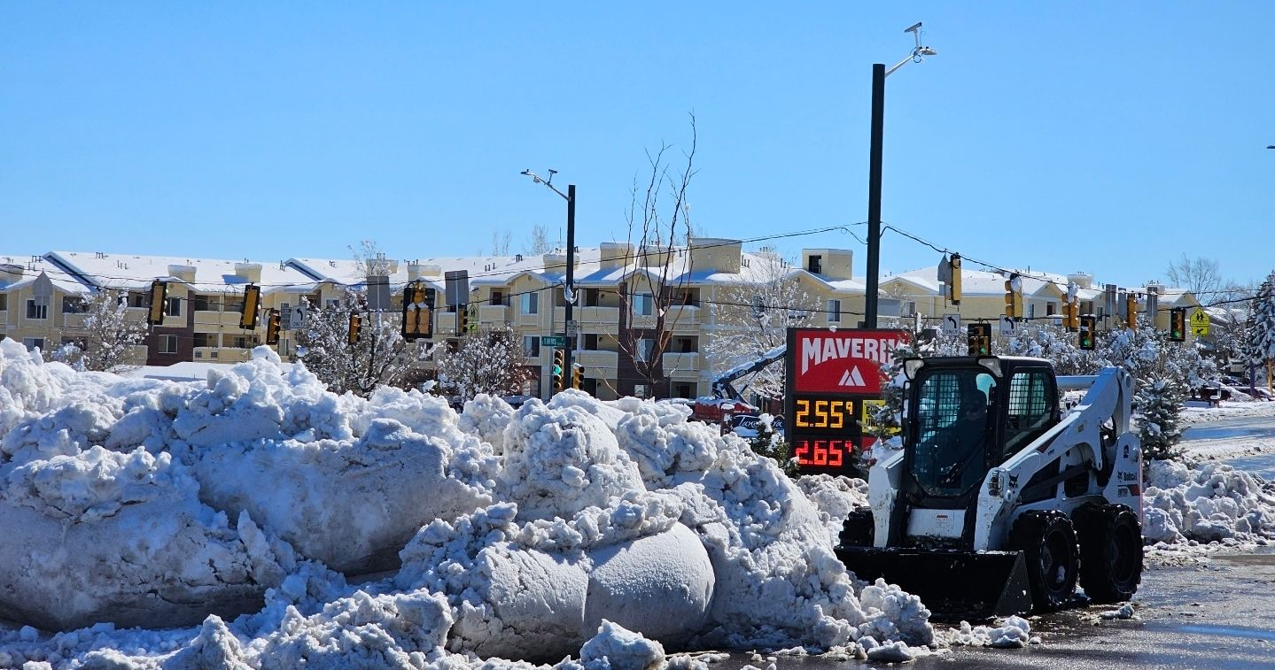 A white skid-steer loader removes snow near a large pile of snow and a gas station sign with a blue sky background.