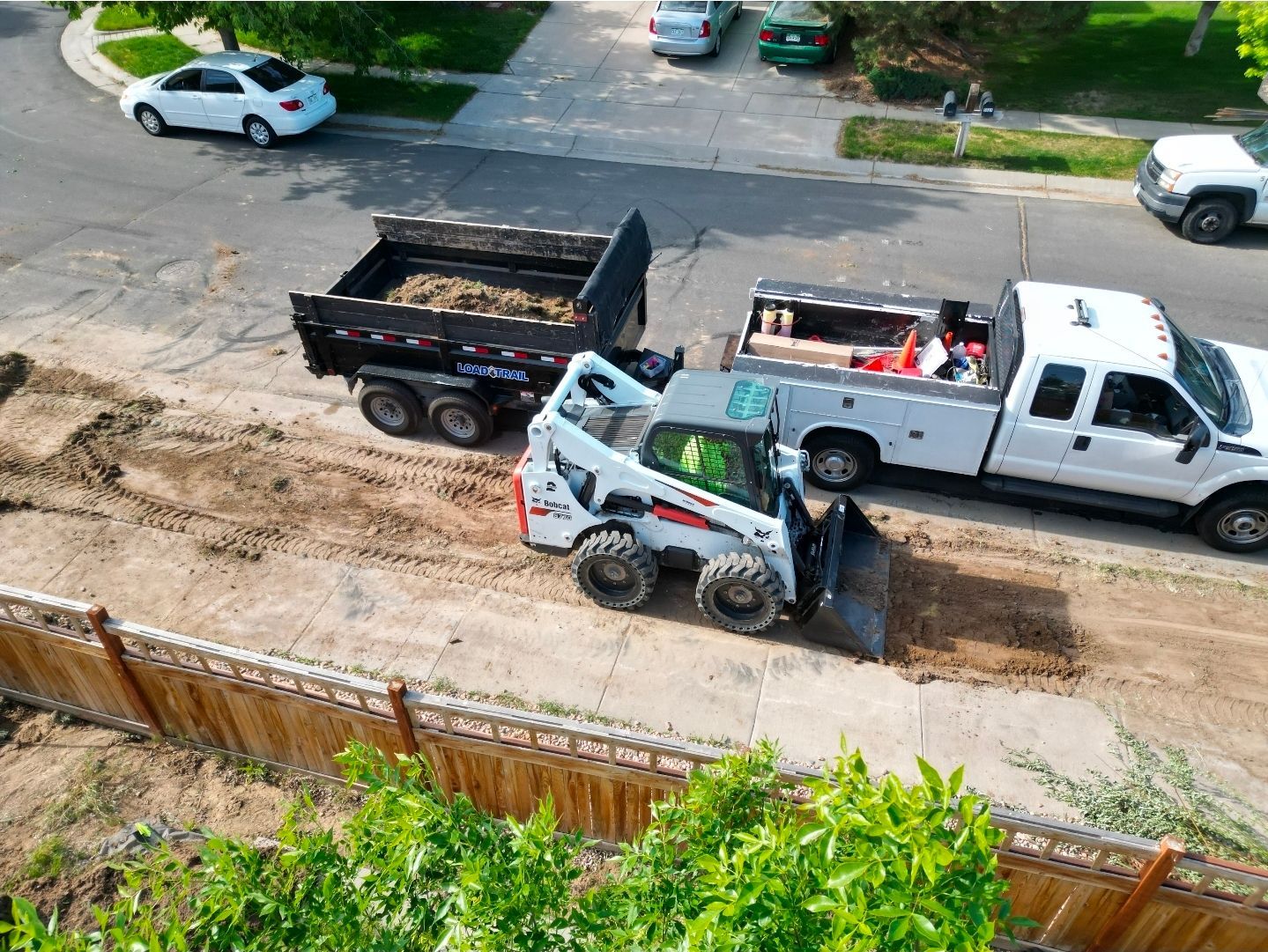A white skid steer loader scoops dirt on a residential street next to a dump trailer and a white pickup truck.