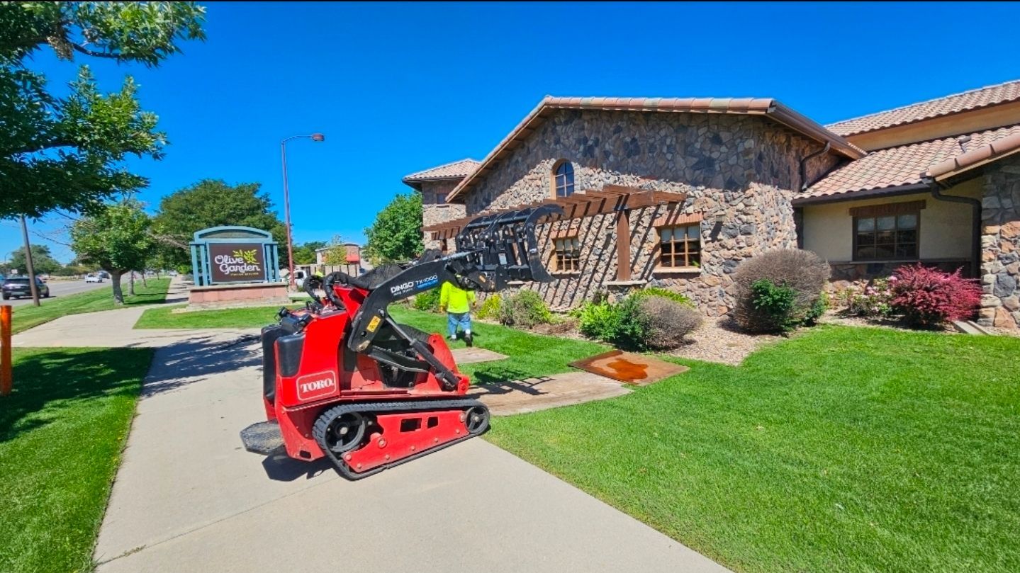 A red skid steer loader parked on a sidewalk in front of a stone building on a sunny day.