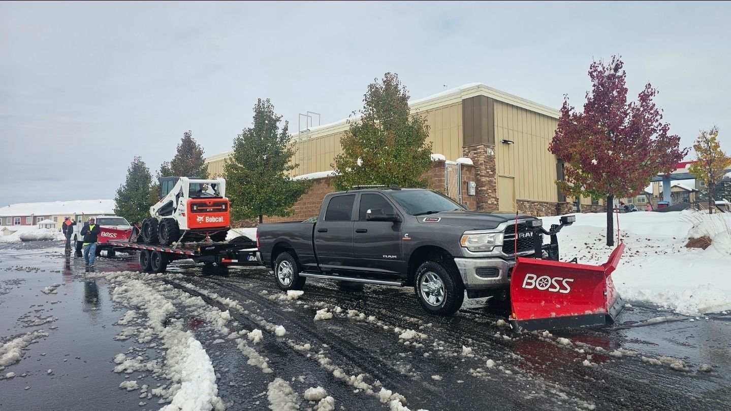 A gray pickup truck with a red Boss snow plow parked on a snowy, slushy lot, towing a trailer with a white Bobcat skid steer.