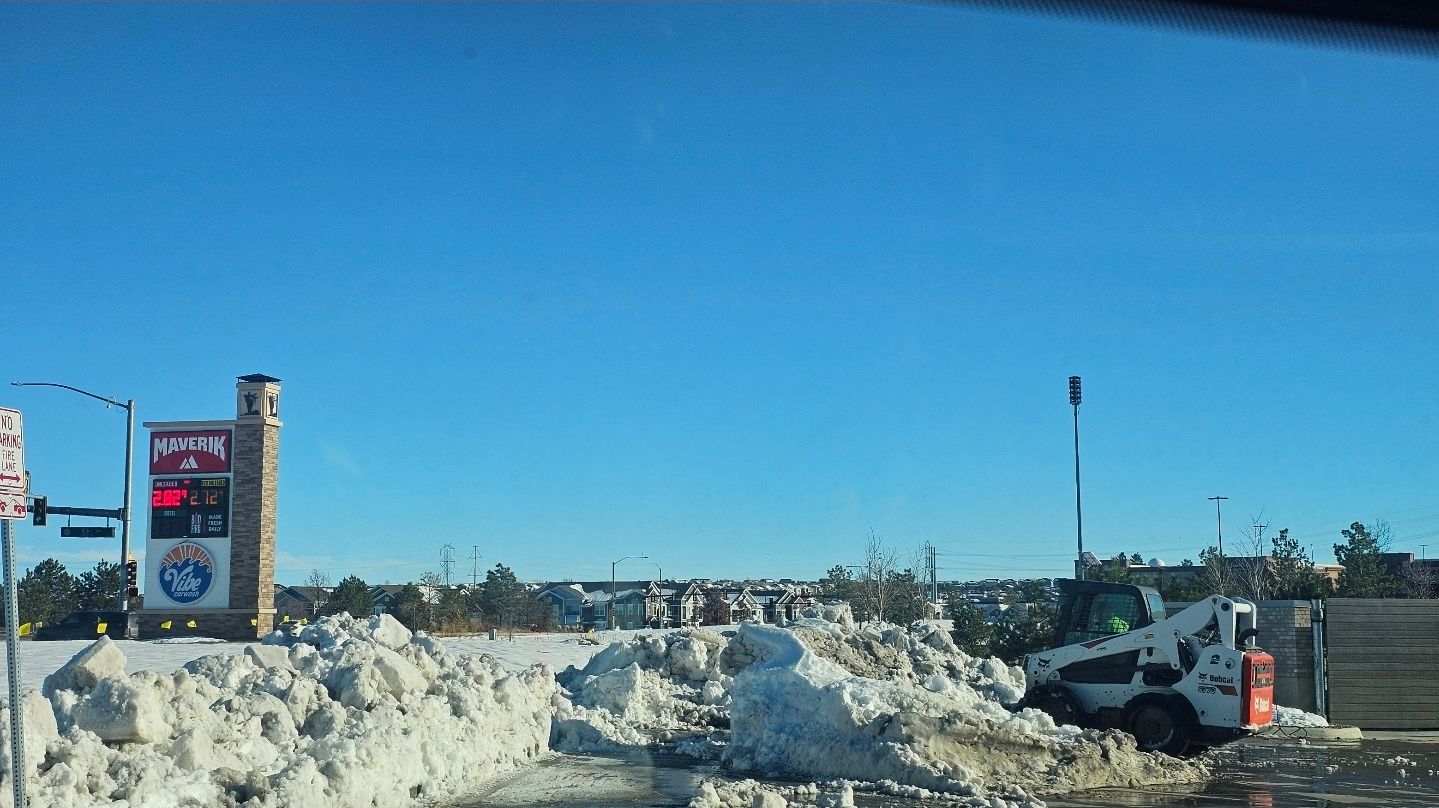 A small white loader clears snow from a parking lot near a commercial stone-pillar sign under a clear blue sky.