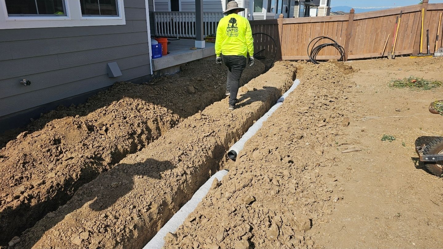 A worker in a high-visibility yellow jacket walks along a trench with a white drainage pipe installed in a dirt yard.