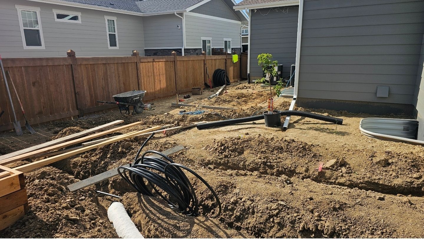 A residential backyard under construction with exposed soil, drainage pipes, a wooden fence, and nearby houses.