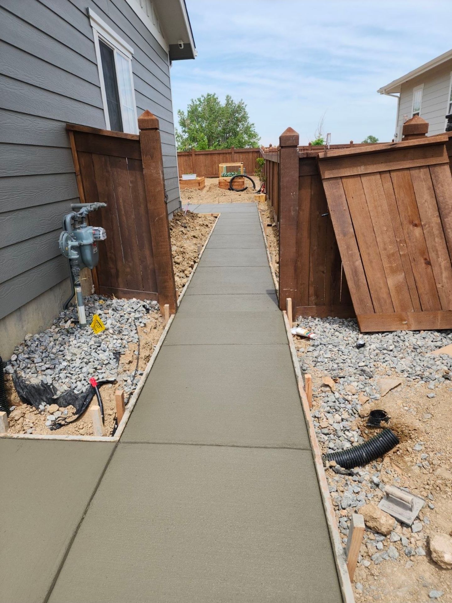 Concrete walkway runs between a house wall and a wooden fence in need of repair.