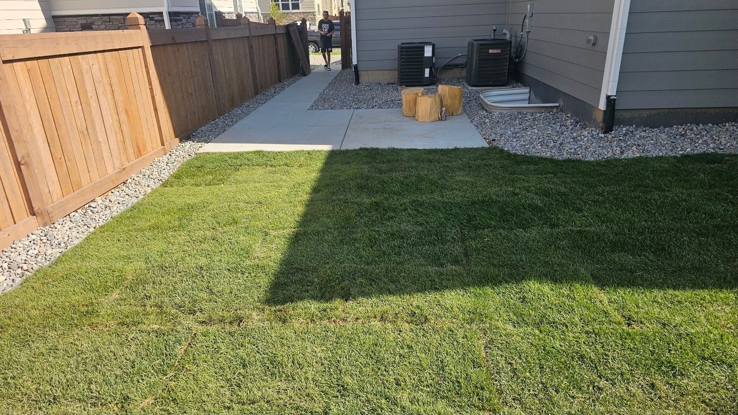 A backyard with a wood fence, a patch of green grass, a concrete walkway, and gravel landscaping near a house exterior.