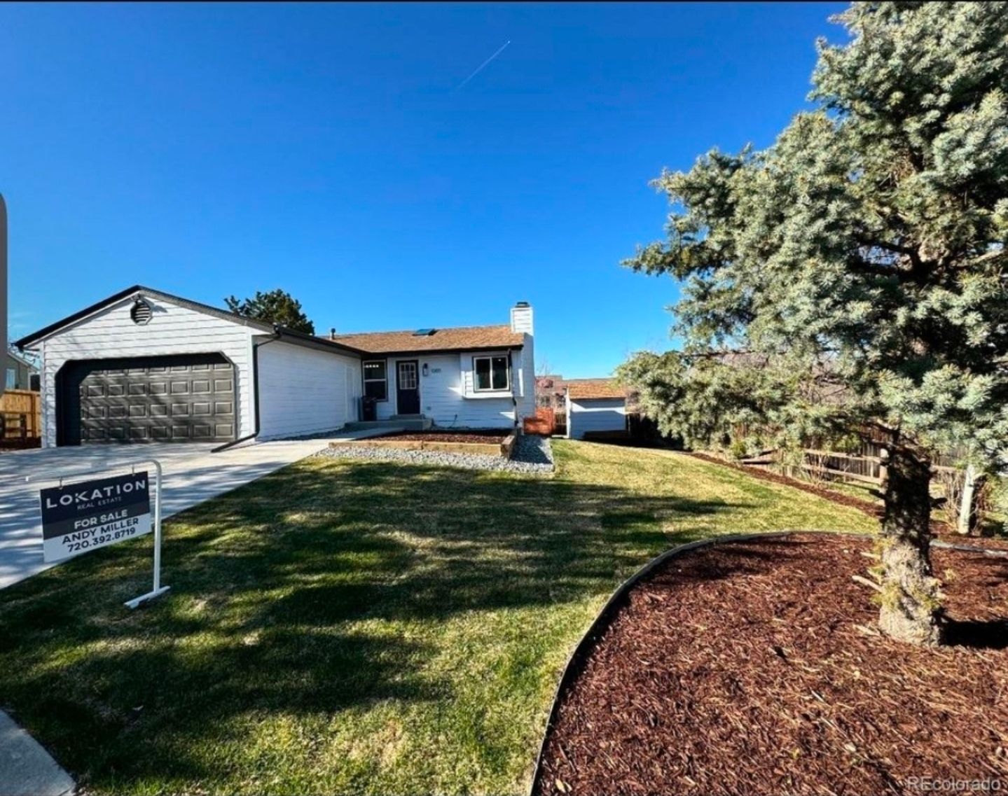 A white, single-story house with a black garage door and a front lawn under a clear blue sky, with a for-sale sign.