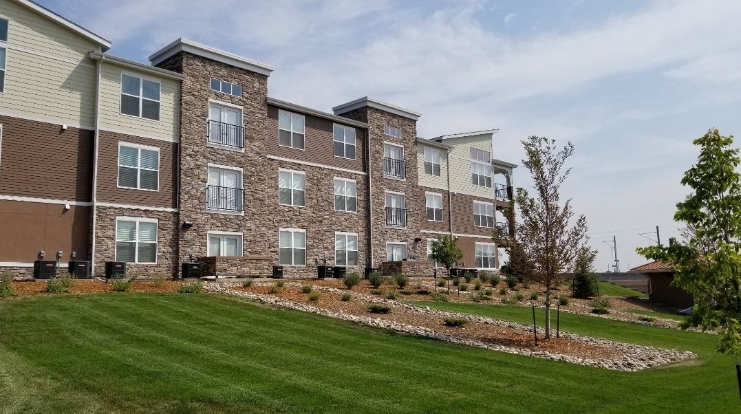 A multi-story apartment building with stone and beige siding facade, set behind a landscaped lawn and rock garden.