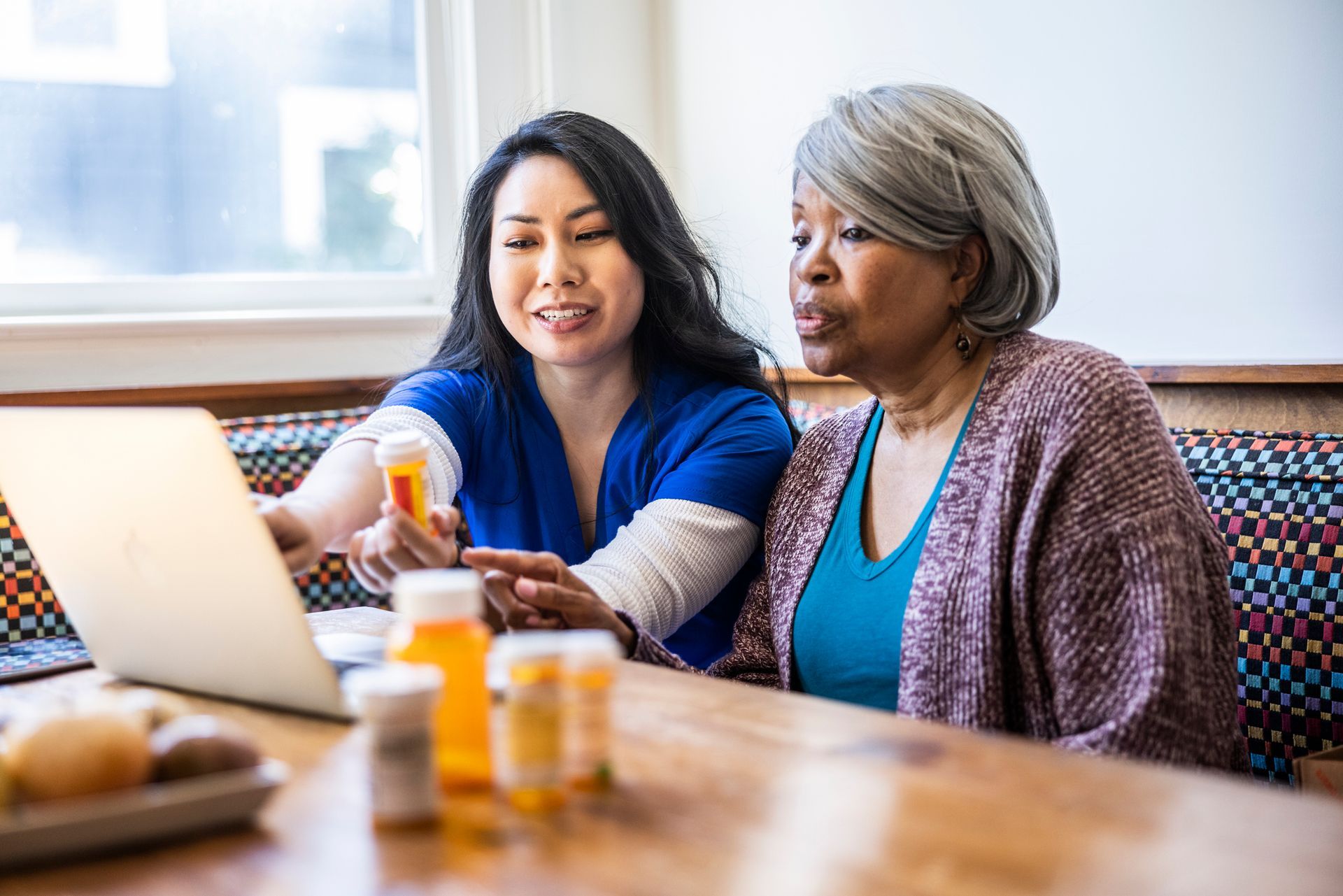 Woman in blue shows pills to another woman, using a laptop at a table.