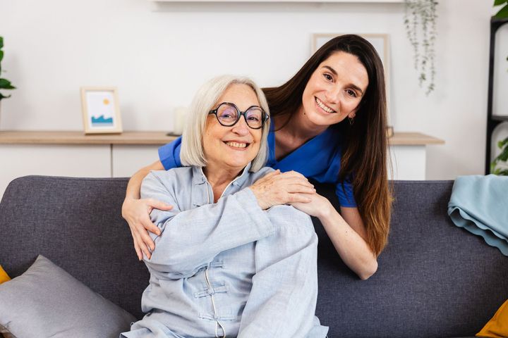 Woman hugs smiling older person sitting on a sofa. Both looking at the camera.