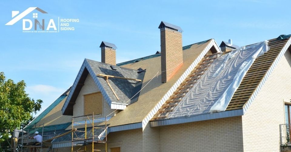 Roofing being installed on a multi-gabled house with two chimneys, sunny day.