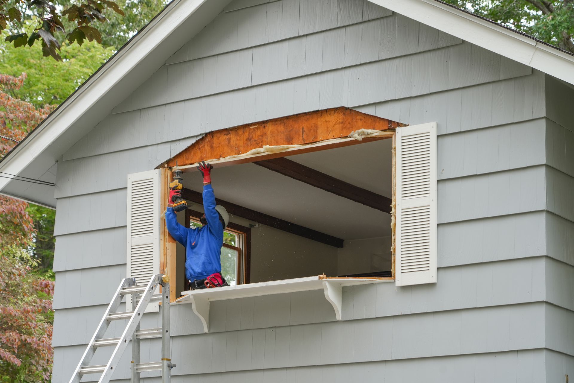 Person using a drill to install a window in a gray house with open shutters; ladder. Person using a drill to install a window in a gray house with open shutters; ladder.