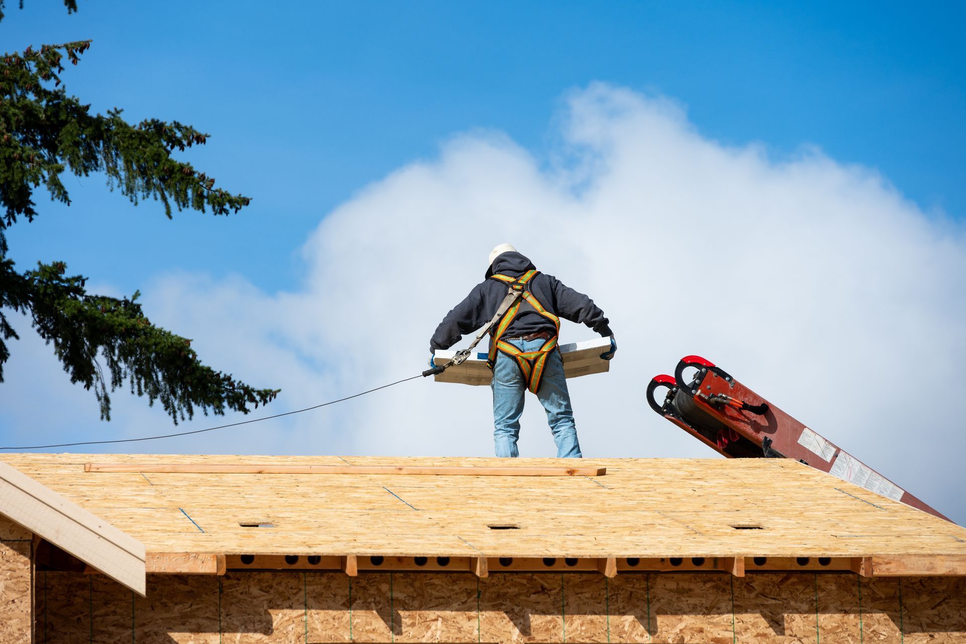 Roofer on a wood roof wearing a harness, operating a lift; blue sky with clouds.