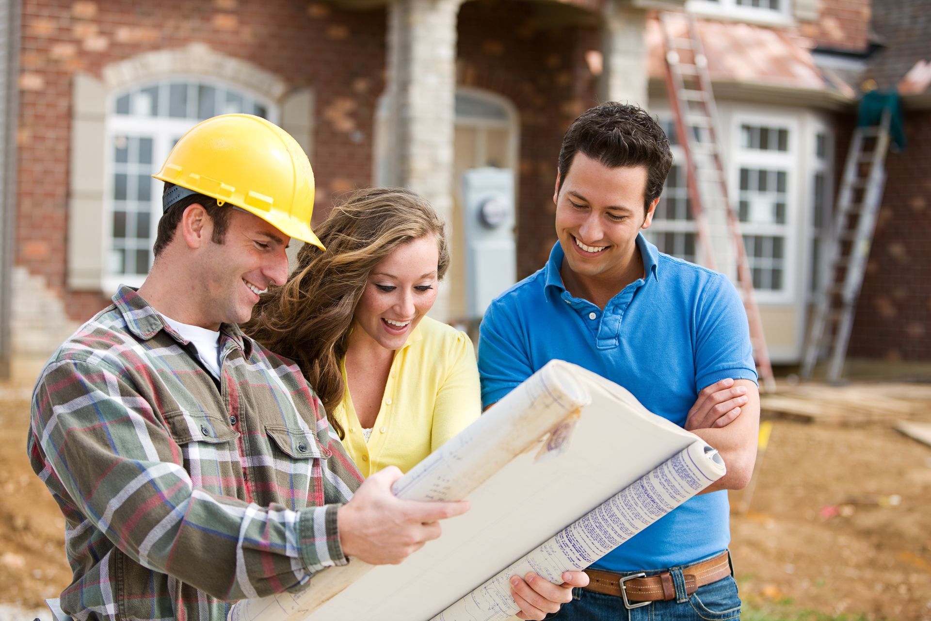 Builder showing blueprints to couple in front of a house under construction.