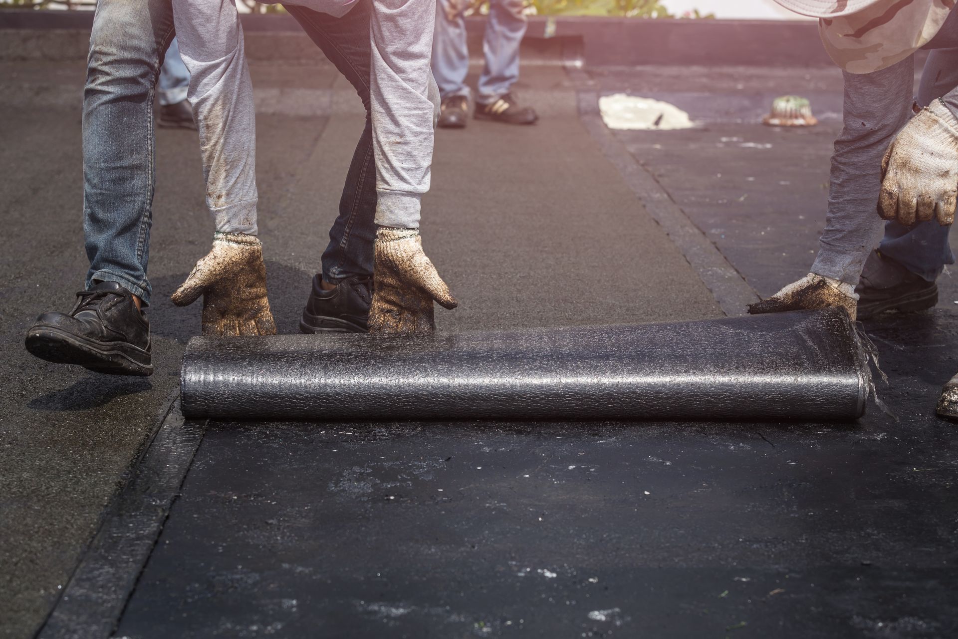 Workers rolling out a dark roofing material on a flat rooftop.