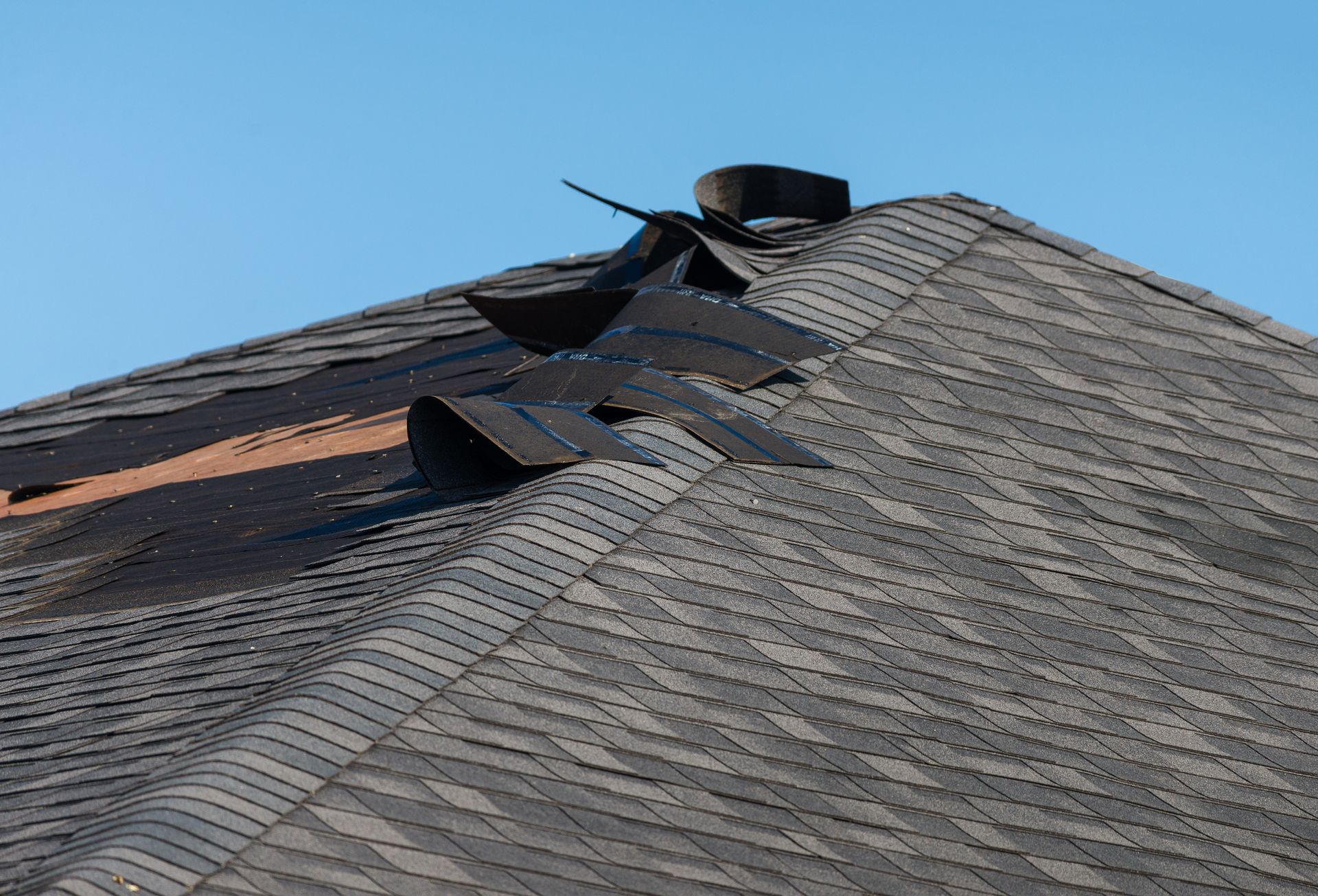 Damaged asphalt shingle roof against a clear blue sky. Torn shingles exposed.