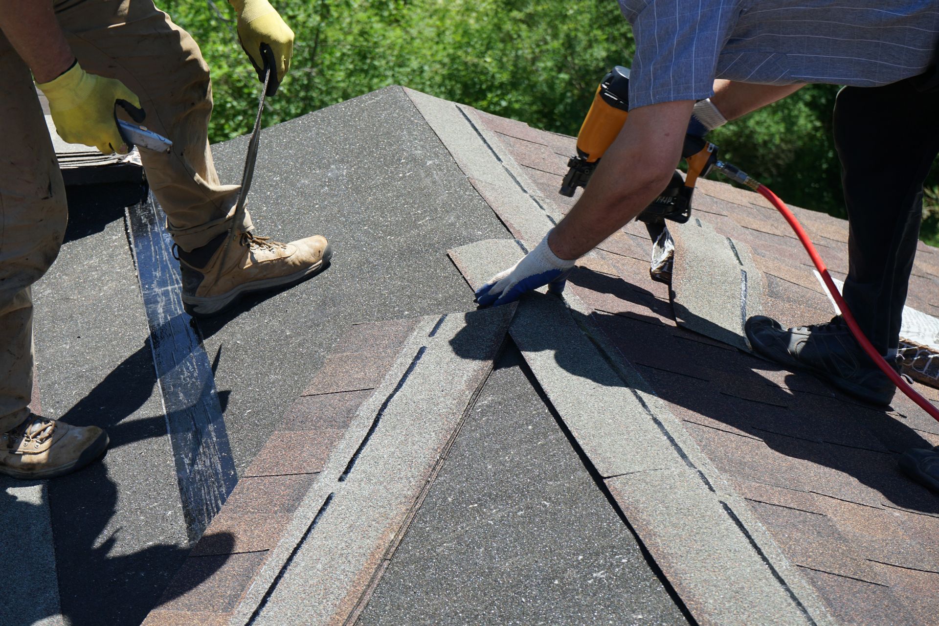 Roofers installing shingles on a roof peak using a nail gun; sunny outdoor setting.
