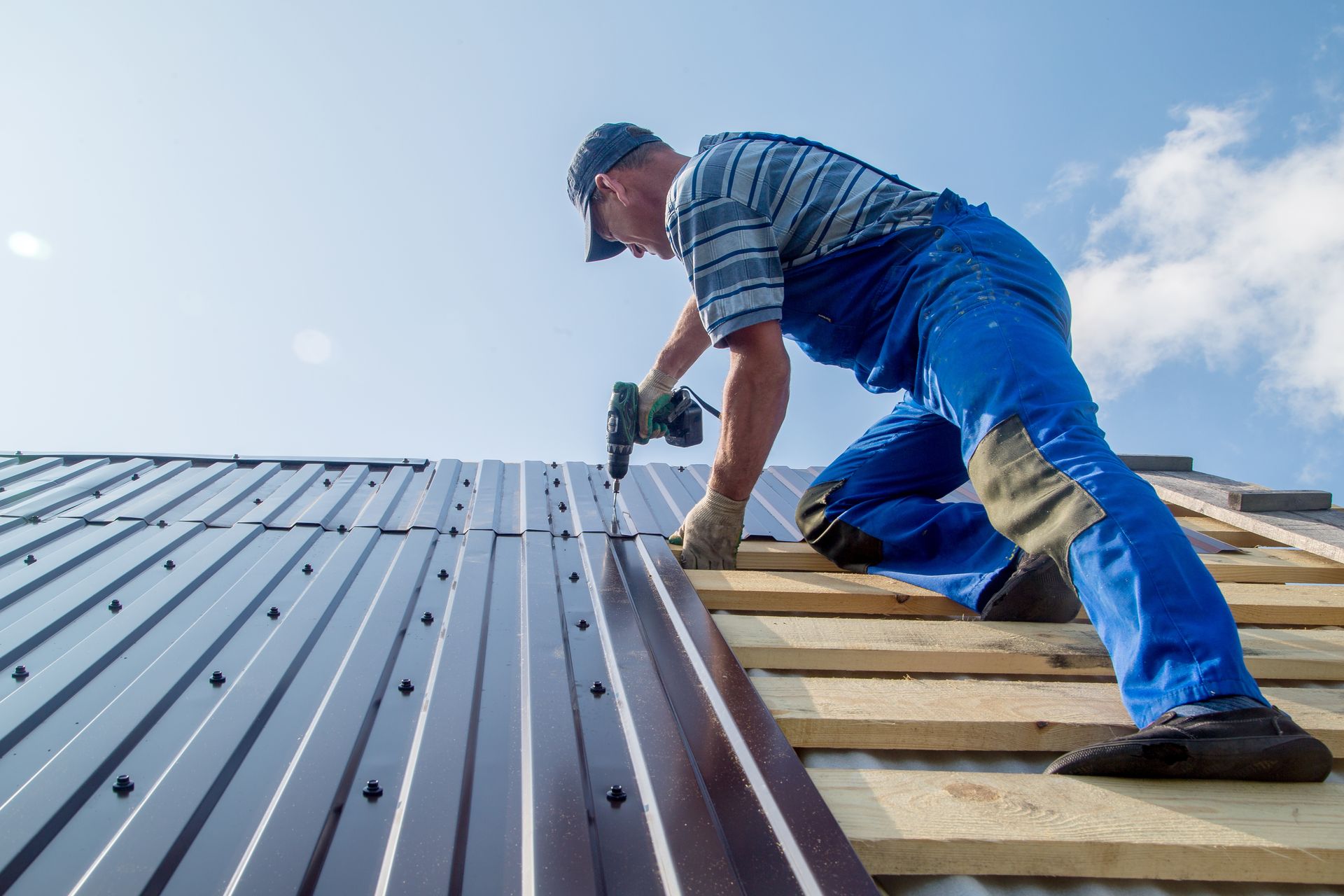 A roofer in blue overalls and cap installing metal roofing on a sunny day.