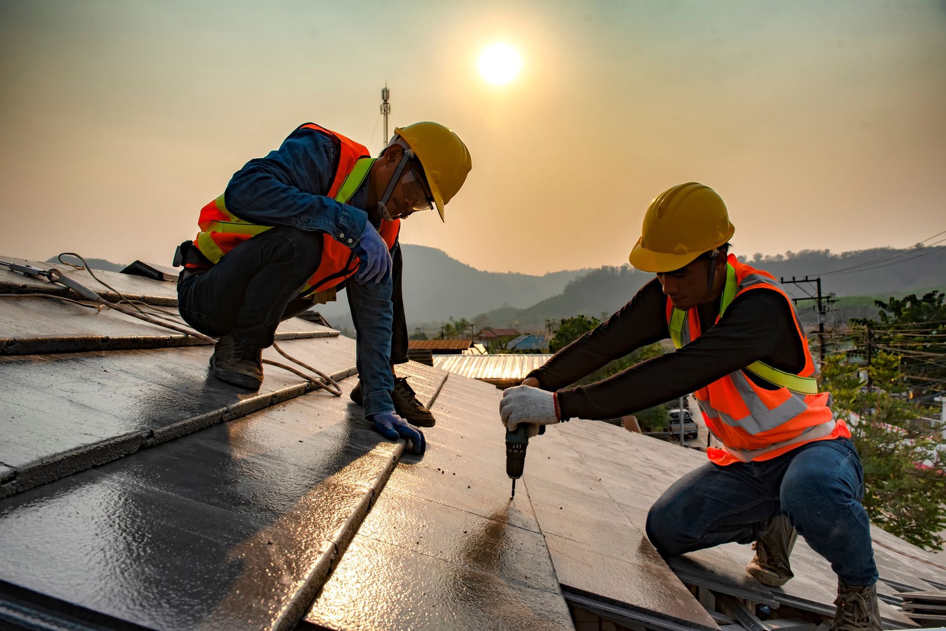 Two workers installing solar panels on a rooftop; both wear helmets and safety vests. Sunlight fills the background.