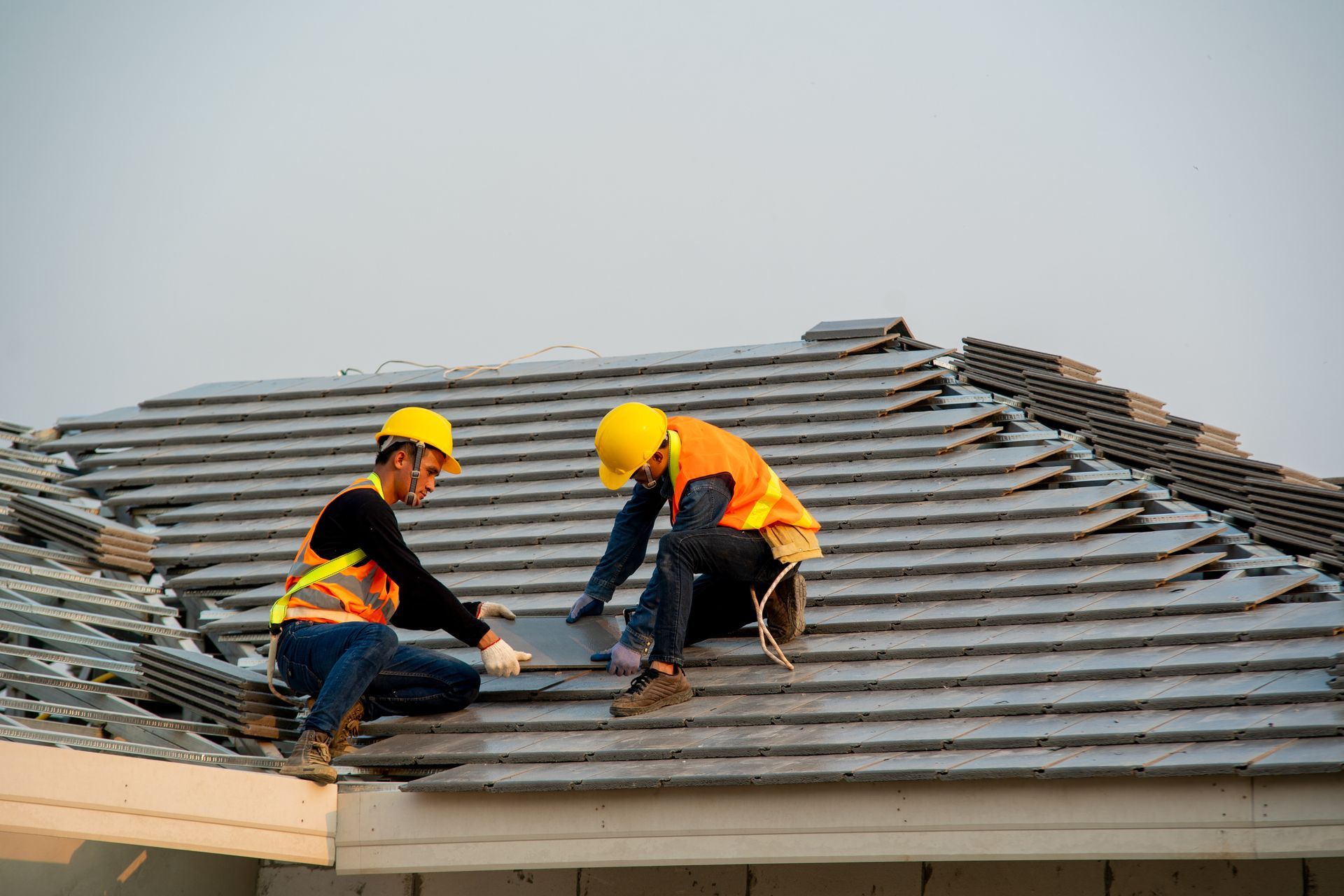 Two roofers in hard hats and vests install gray tiles on a roof.