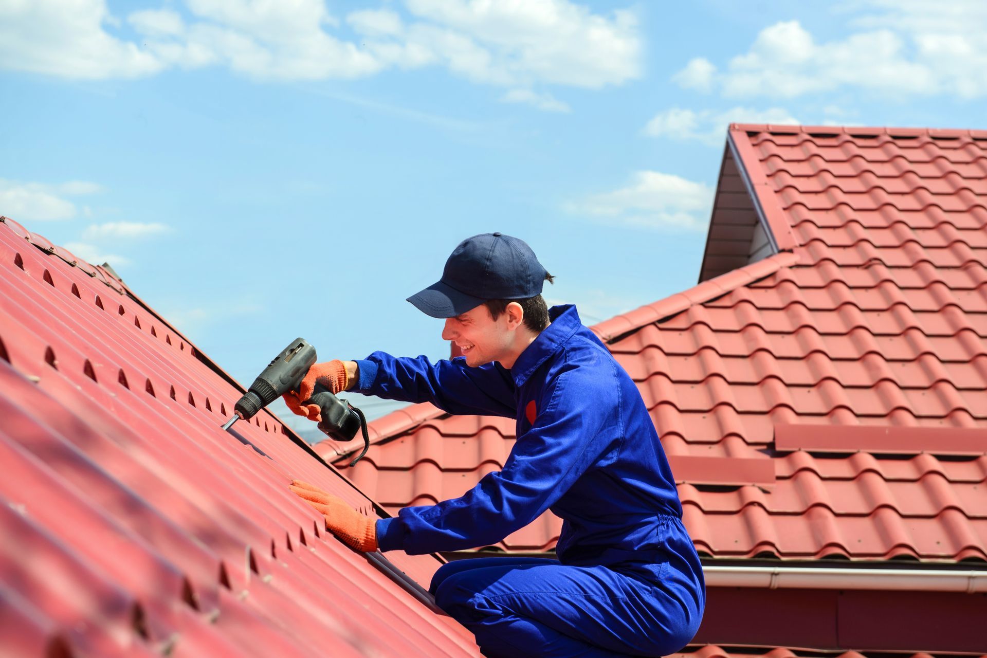 Roofer in blue jumpsuit and cap uses drill on red tile roof against blue sky.