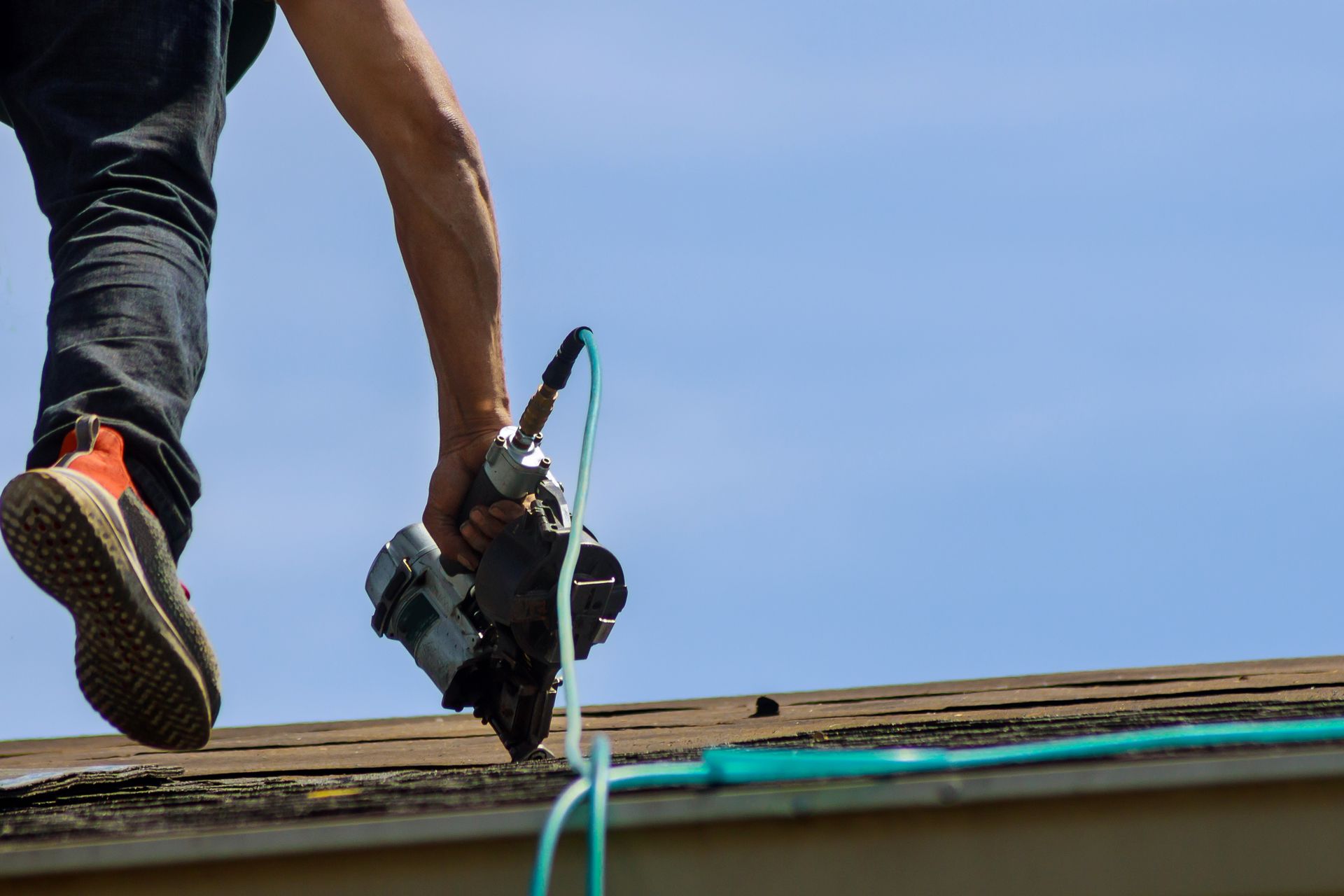 Roofer on a roof, holding a nail gun, blue sky in background.