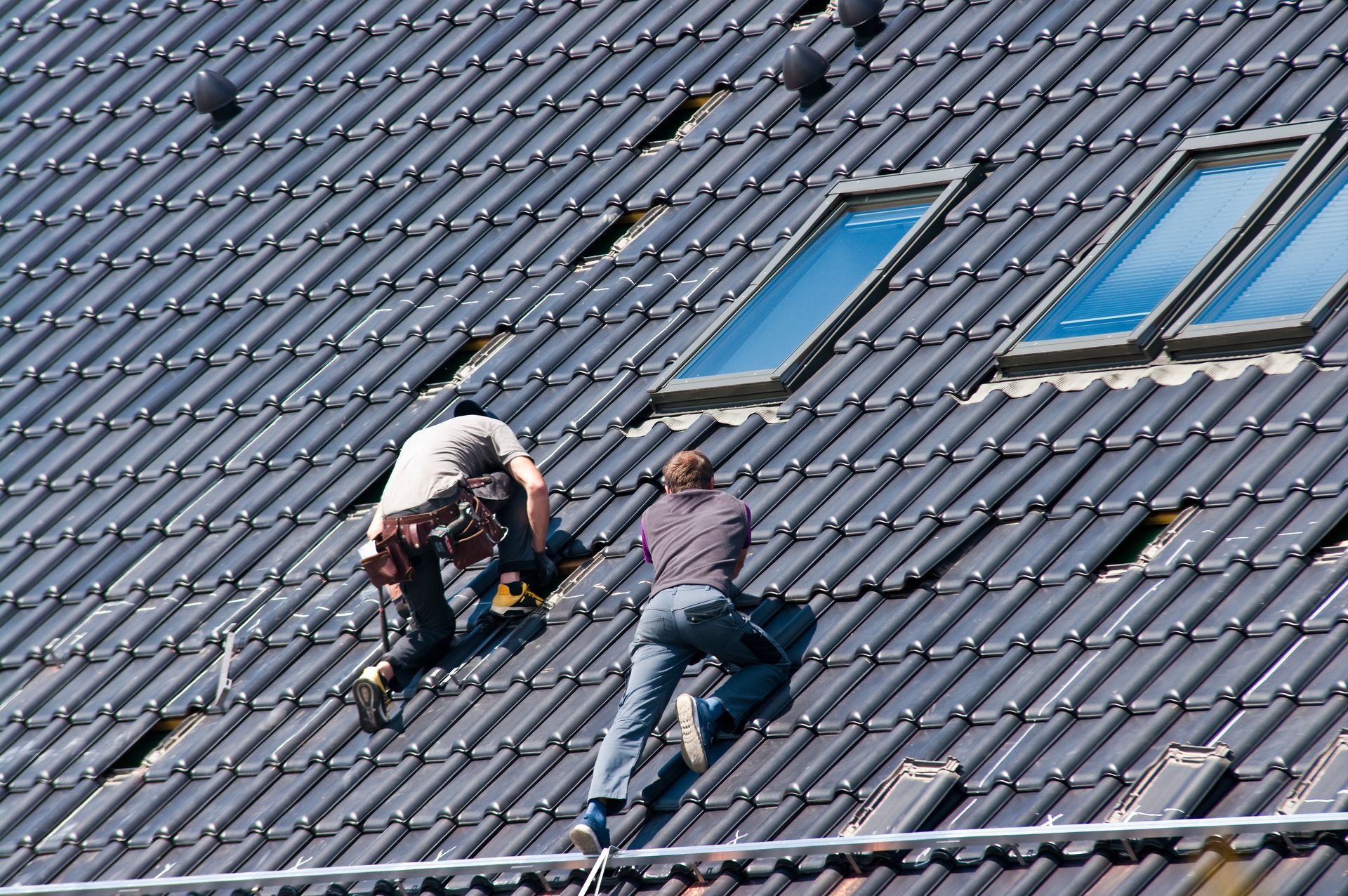 Two roofers on a tiled roof near skylights; one uses a tool.