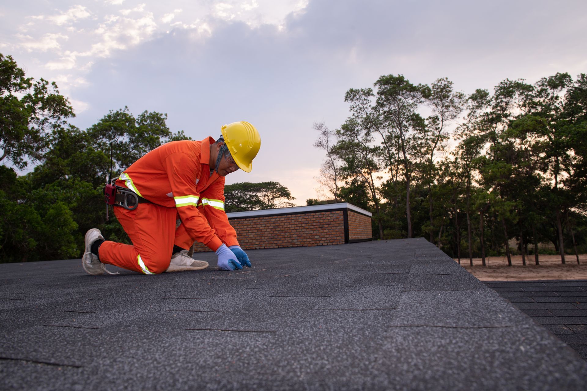 Roofer kneels on a dark shingled roof, wearing orange overalls and a yellow hard hat. Trees and cloudy sky in the background.