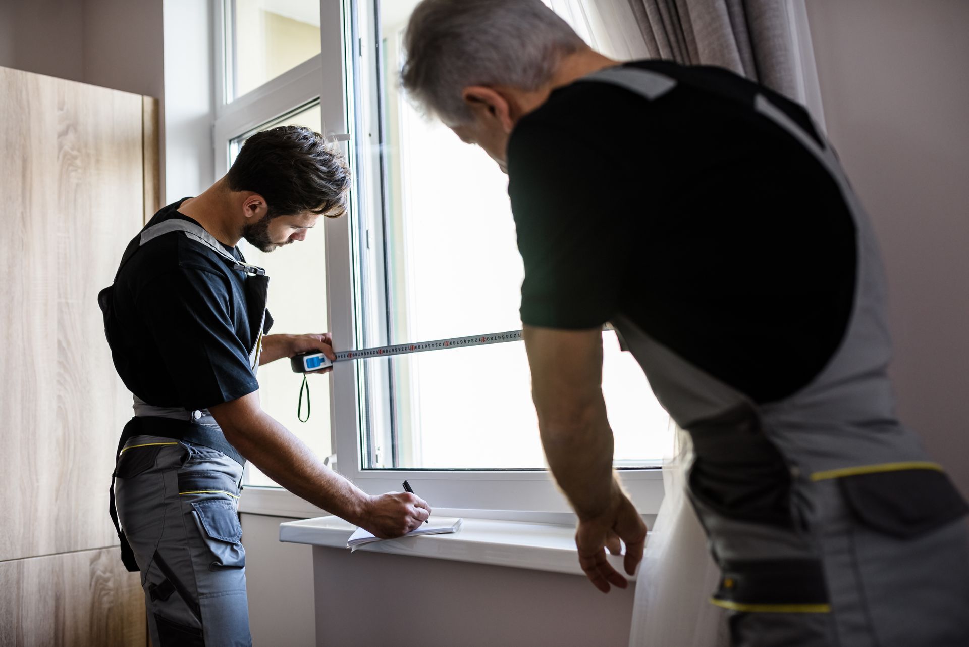 Two people measuring a window for installation, one marking measurements, indoors. Two people measuring a window for installation, one marking measurements, indoors.