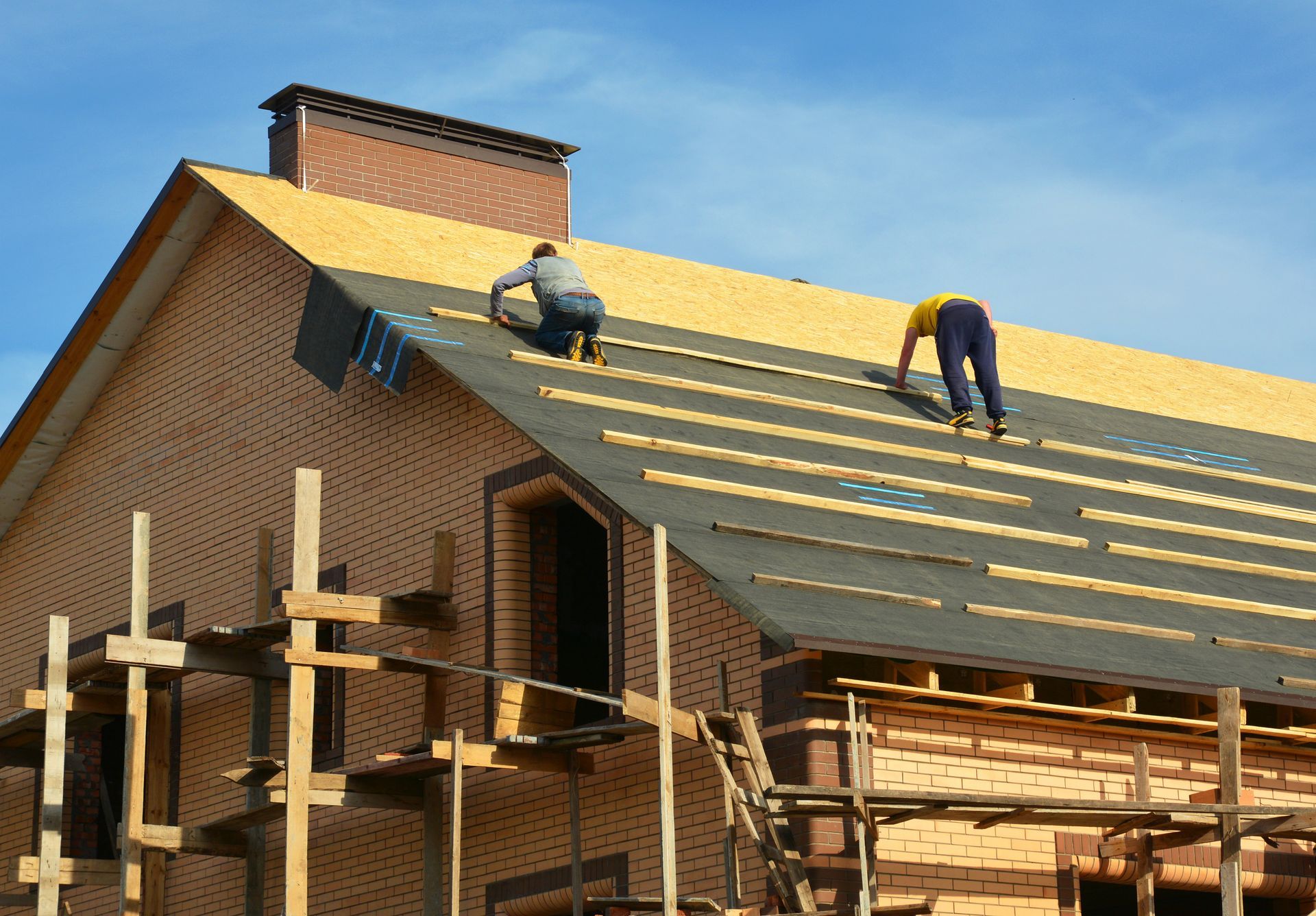 Two workers installing roof shingles on a brick house under construction.