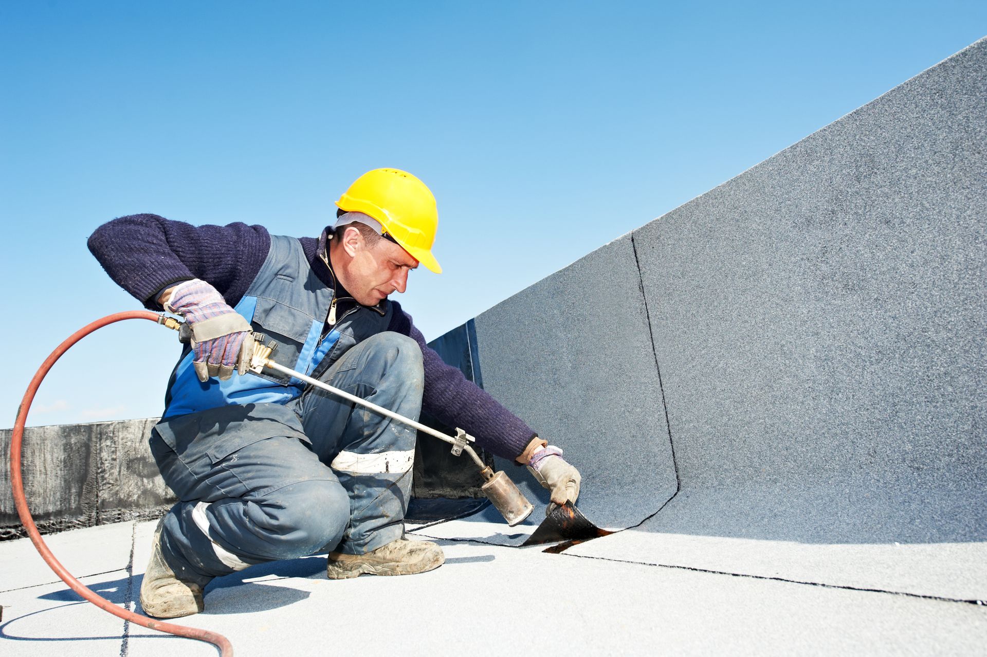 Roofer using a torch to seal seams on a flat roof, kneeling in the sunlight.