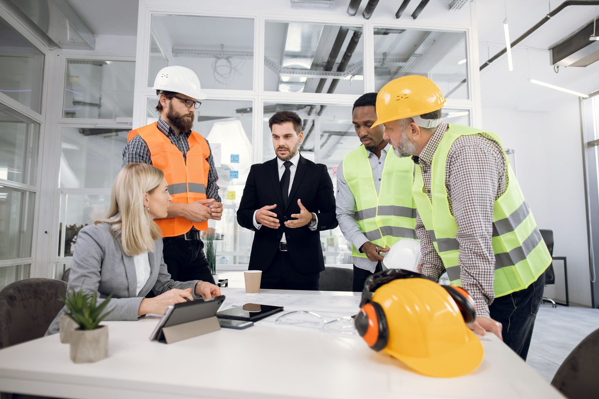 Group of people in construction vests and suits around a table, looking at plans. Office setting.