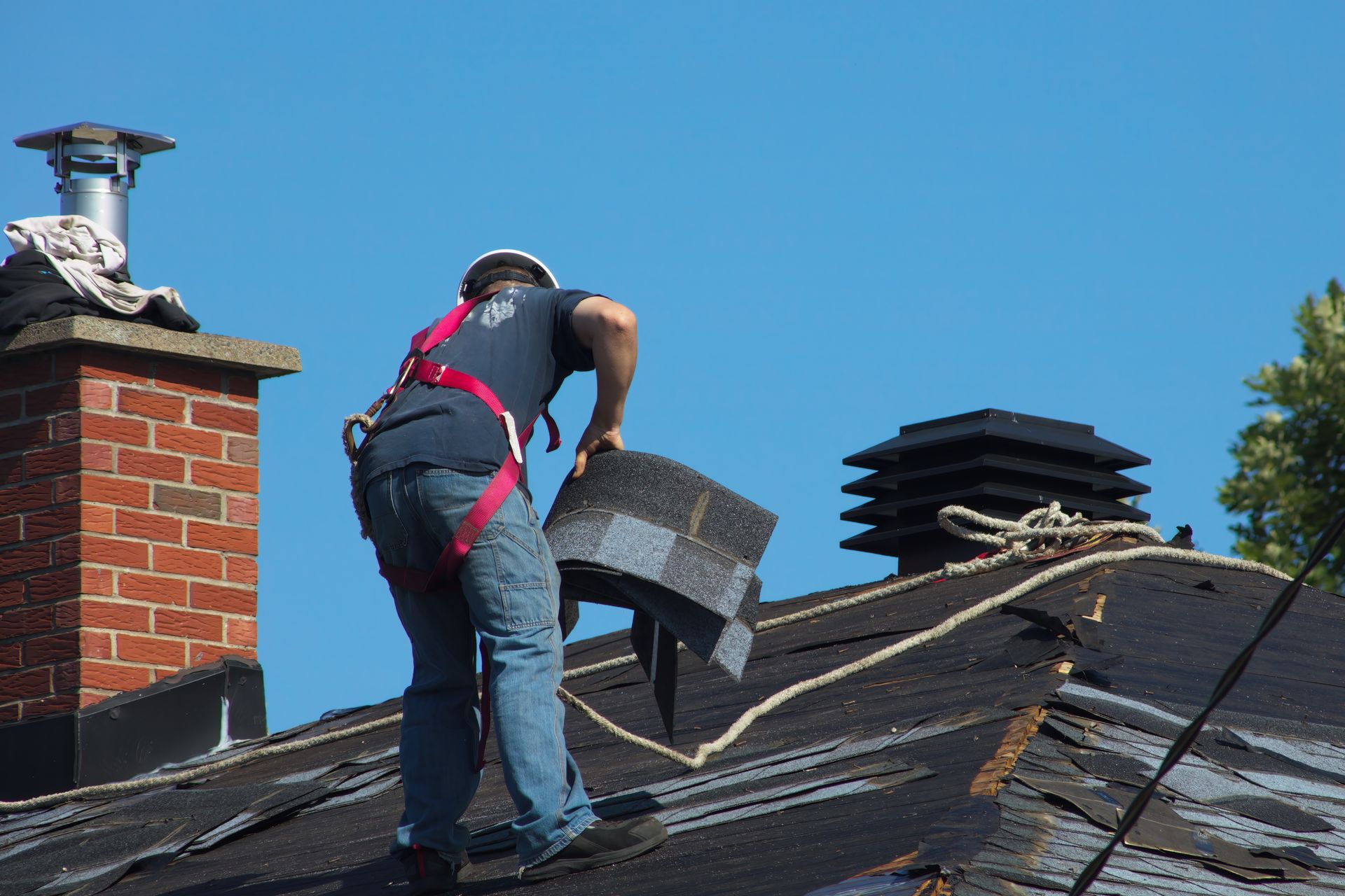Roofer on a dark roof replacing shingles, wearing a safety harness near a brick chimney.