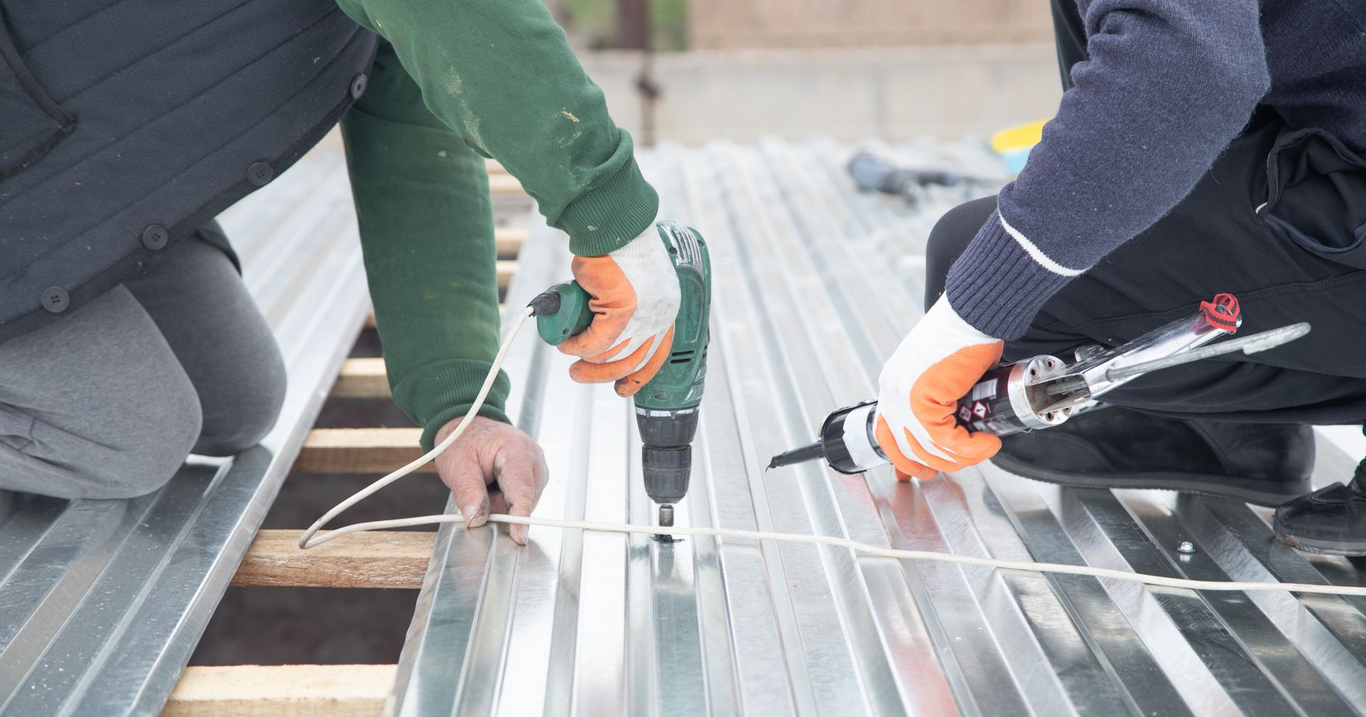 Two people install metal roofing using a drill and caulk gun.