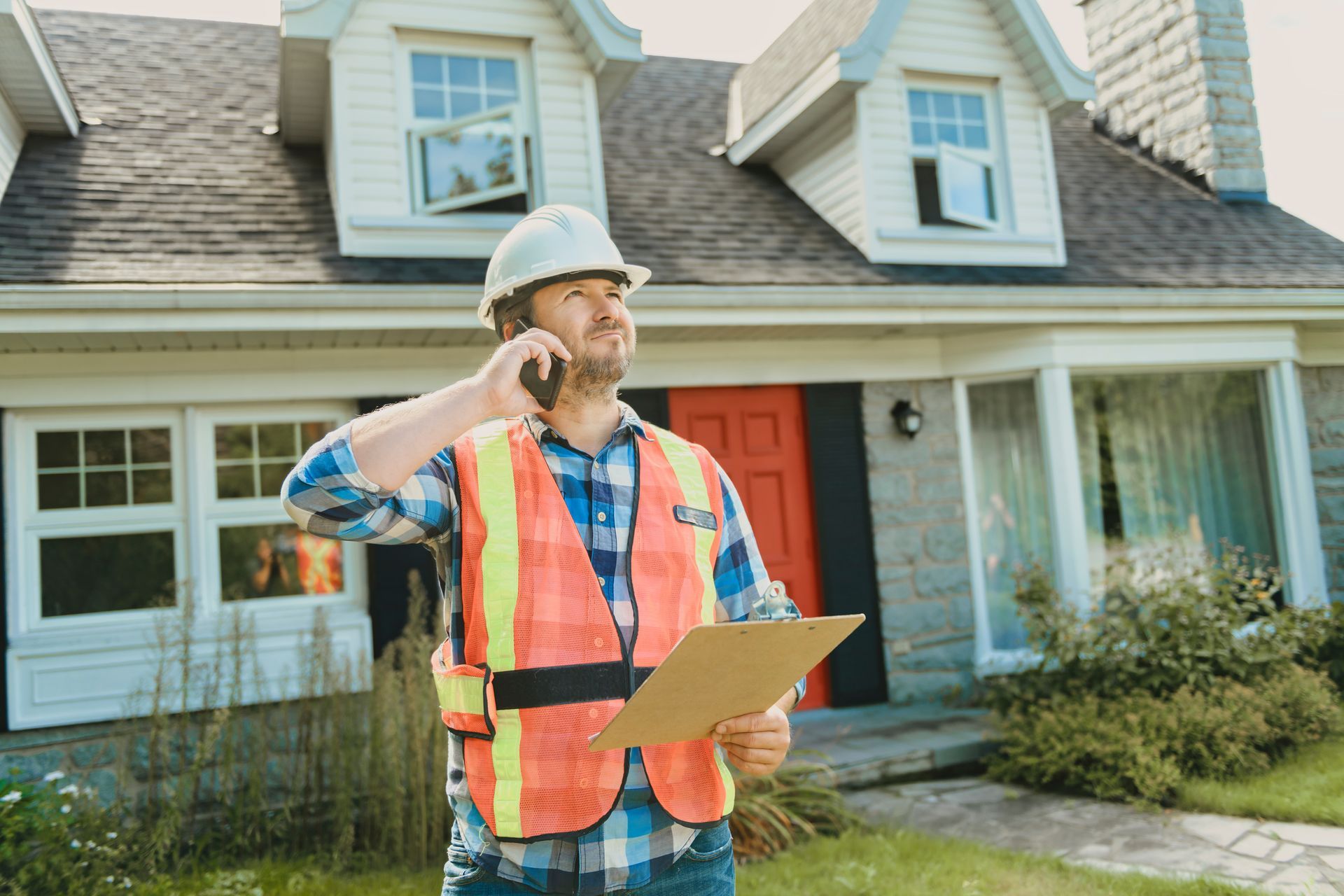 Construction worker in hard hat and vest talks on phone outside house, holding a clipboard.