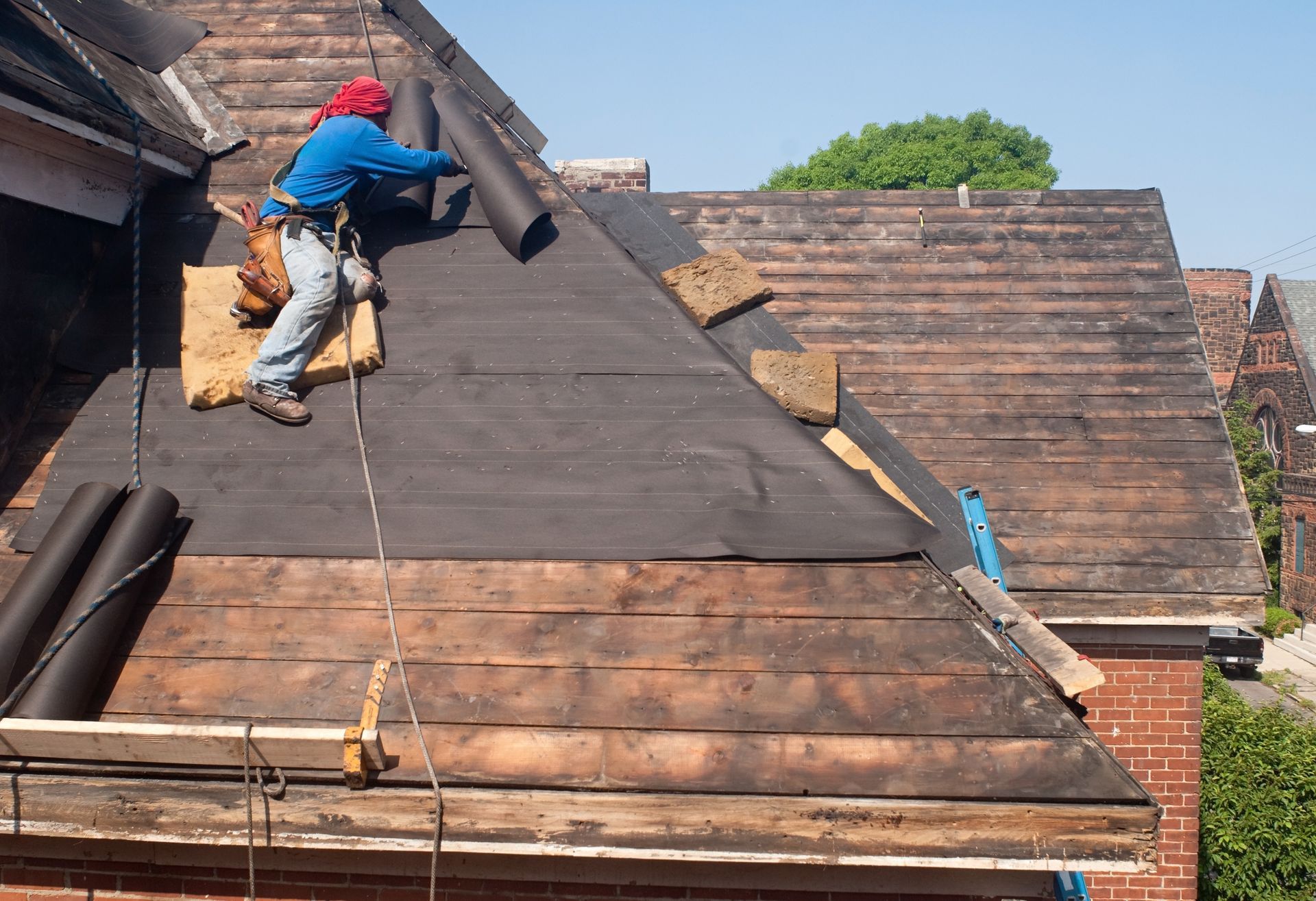 Roofer on a roof, installing black underlayment. Brick building, blue sky.