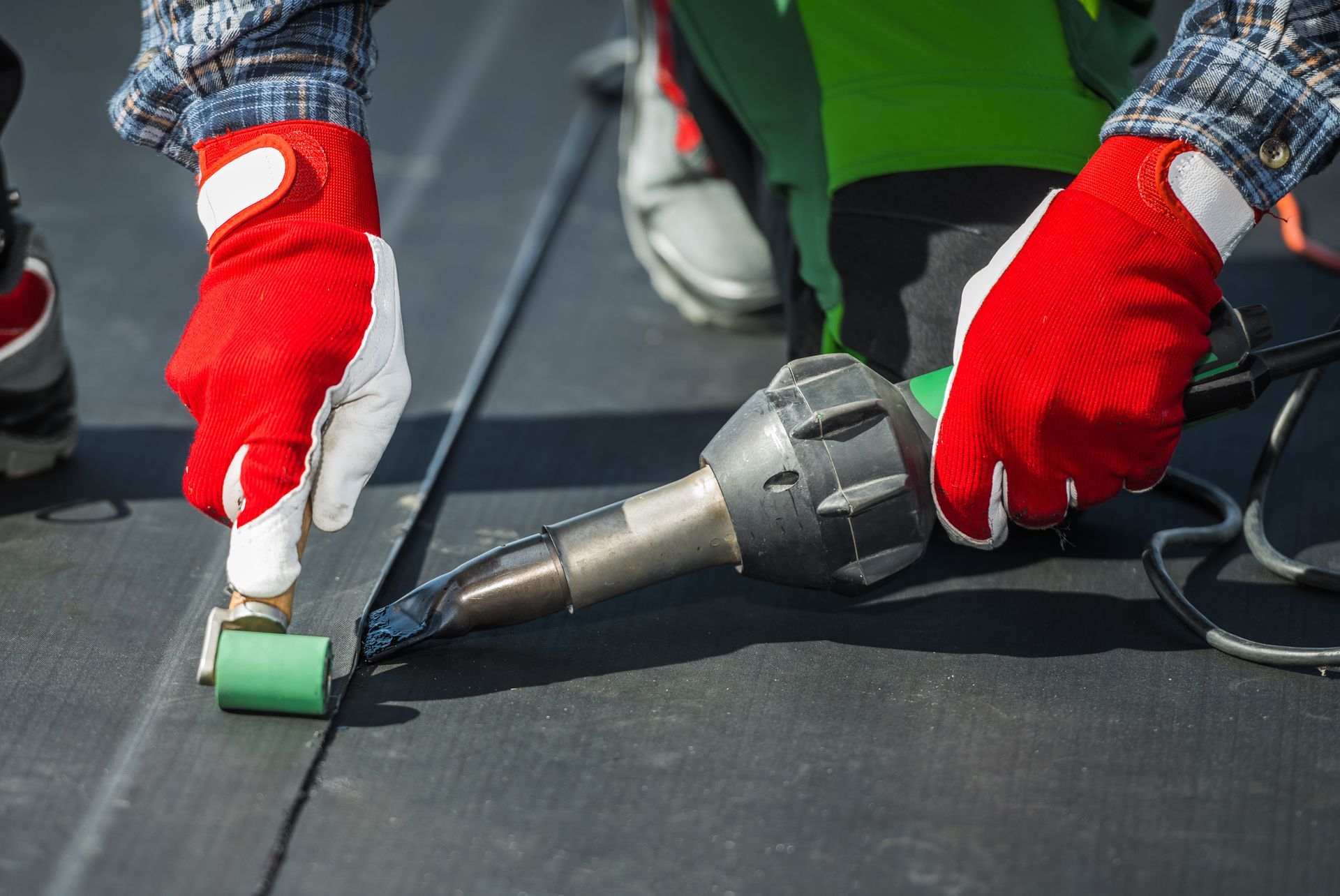 Roofer using a heat gun and roller to seal a black roof seam. Red and white gloves.