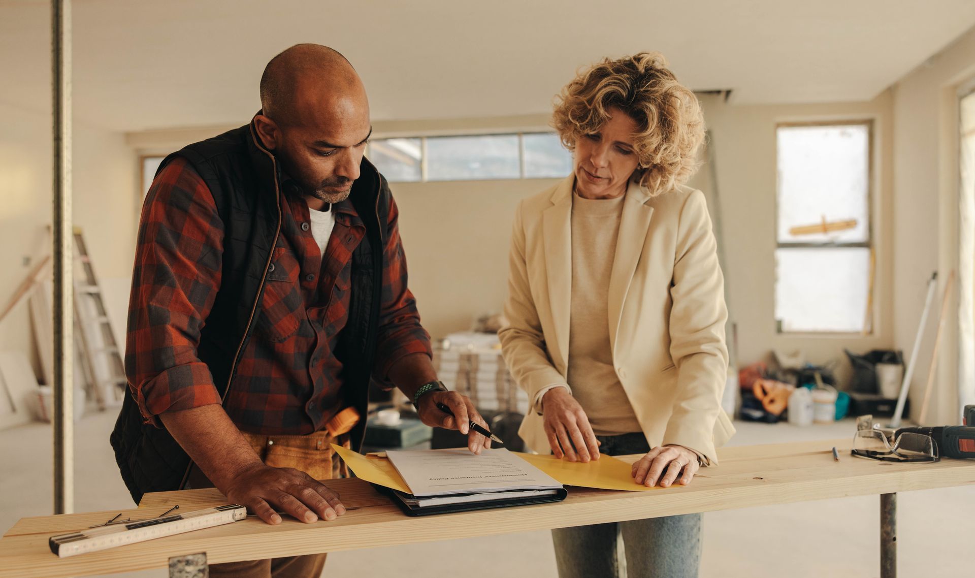 A person with a tool belt and another person review blueprints on a wooden surface in a room under construction.