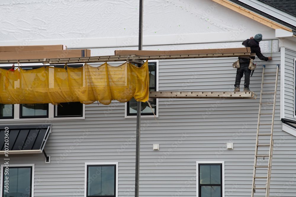 Construction worker on ladder installing siding on a multi-story building. Yellow safety netting and scaffolding are present.