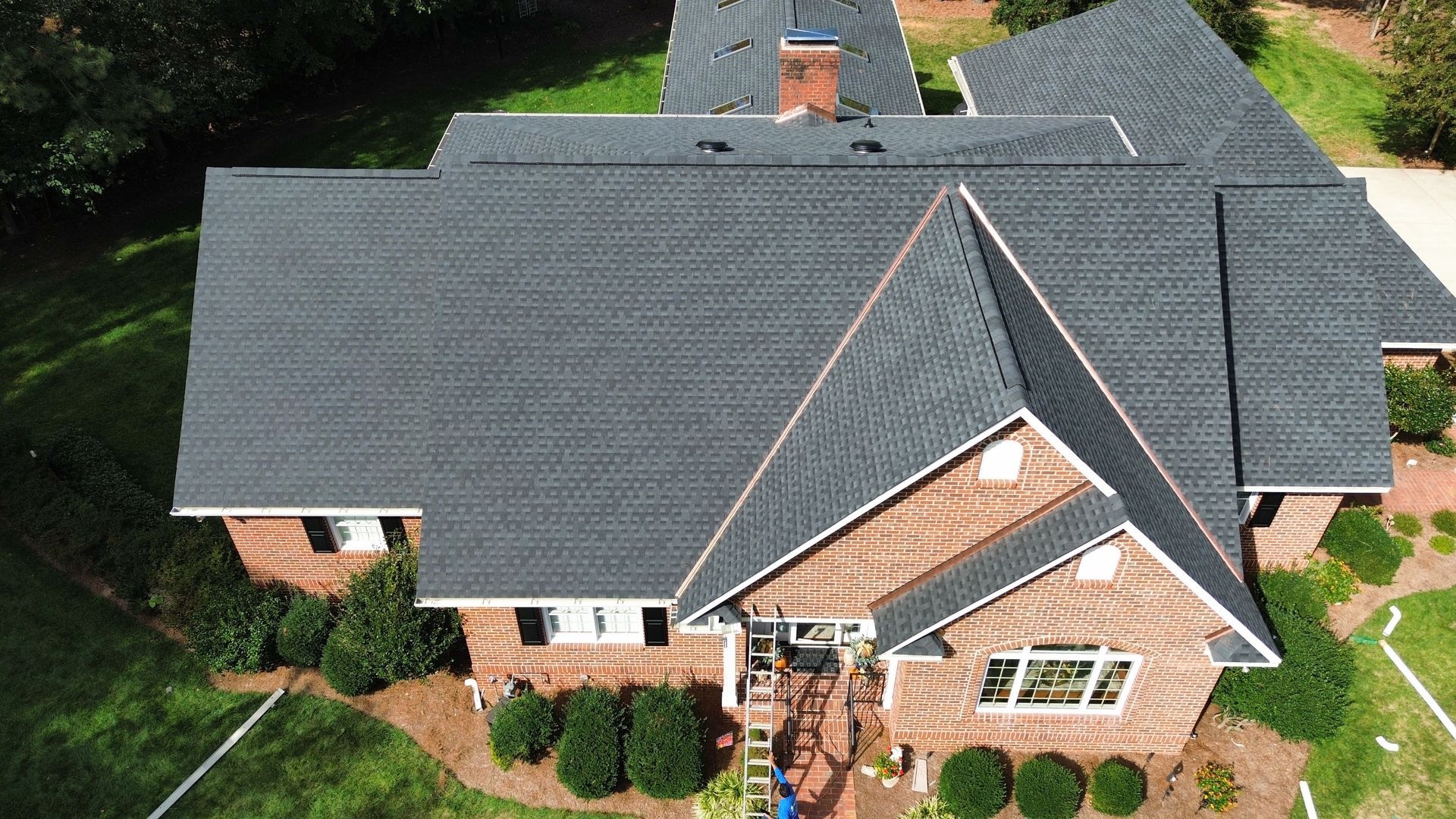 A house with a dark gray shingled roof, brown brick exterior, and green lawn.