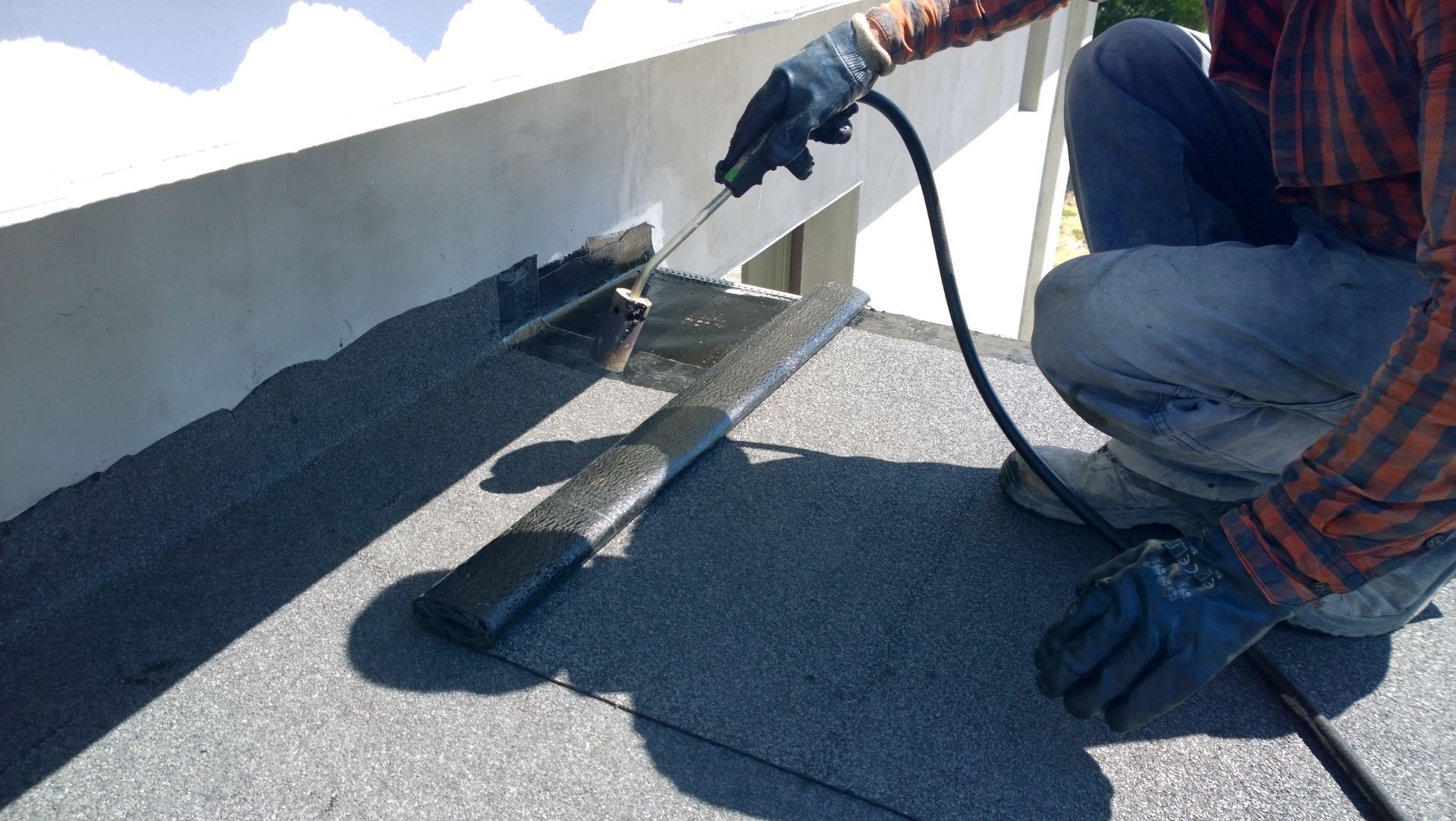 Person using a torch to apply roofing material on a flat roof next to a white wall.