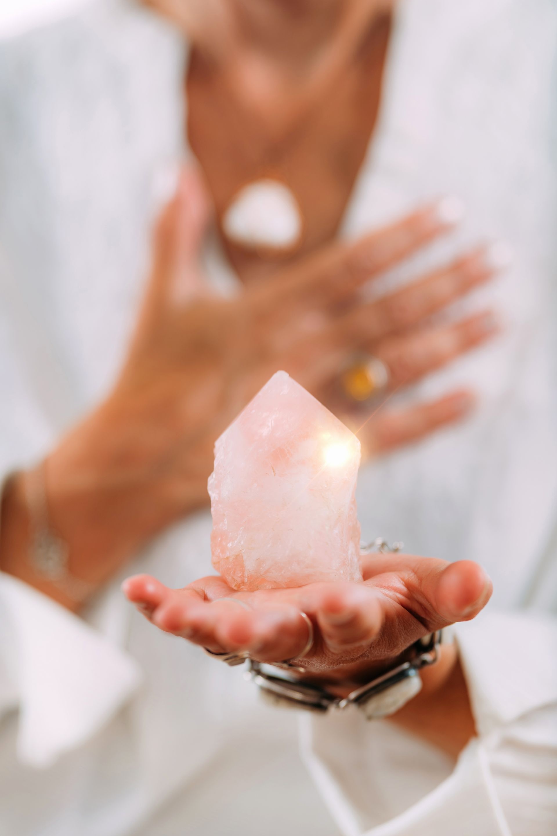 Person holding a glowing pink crystal in their hand, another hand resting on their chest.