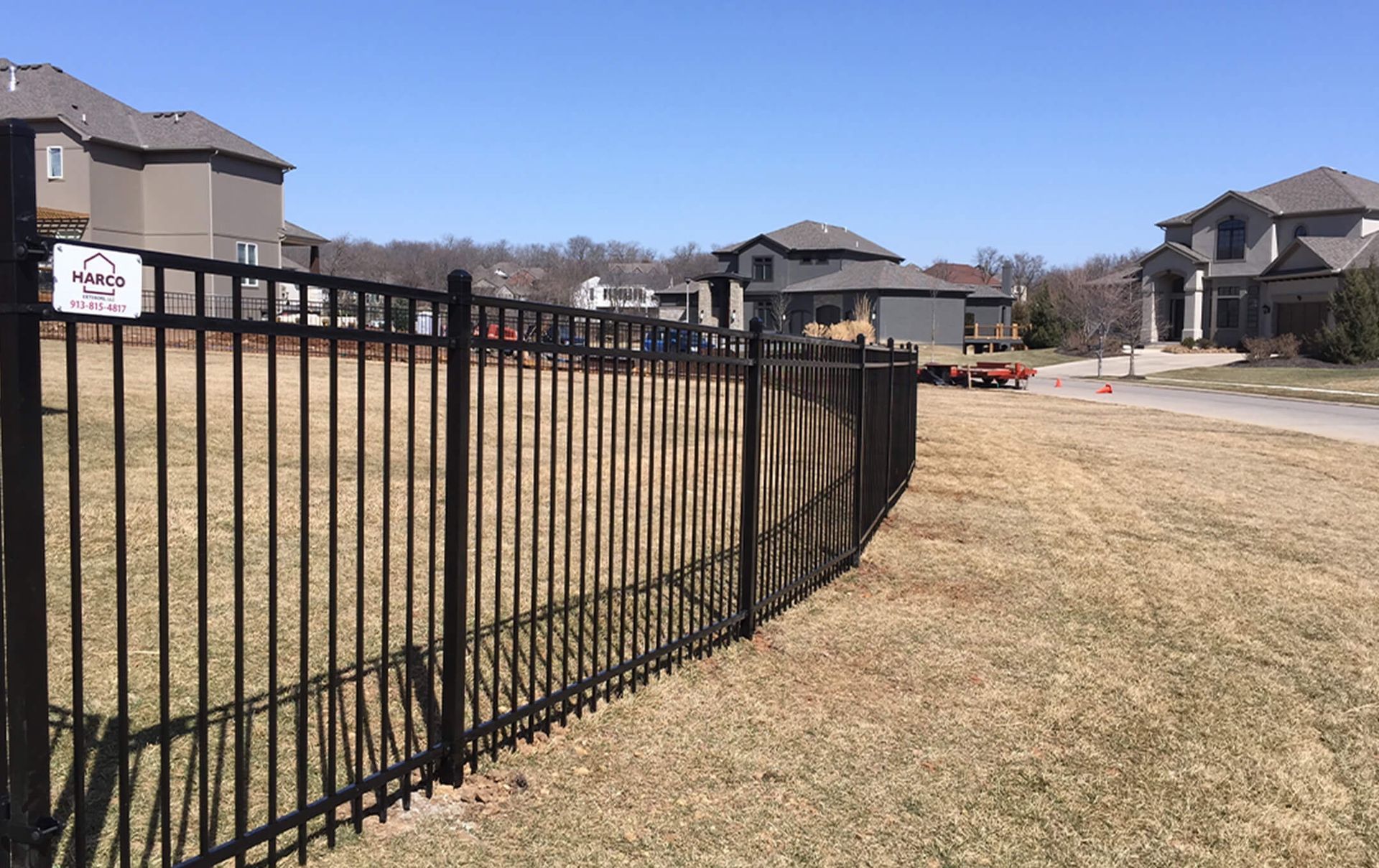 A black metal fence surrounds a grassy field with houses in the background.