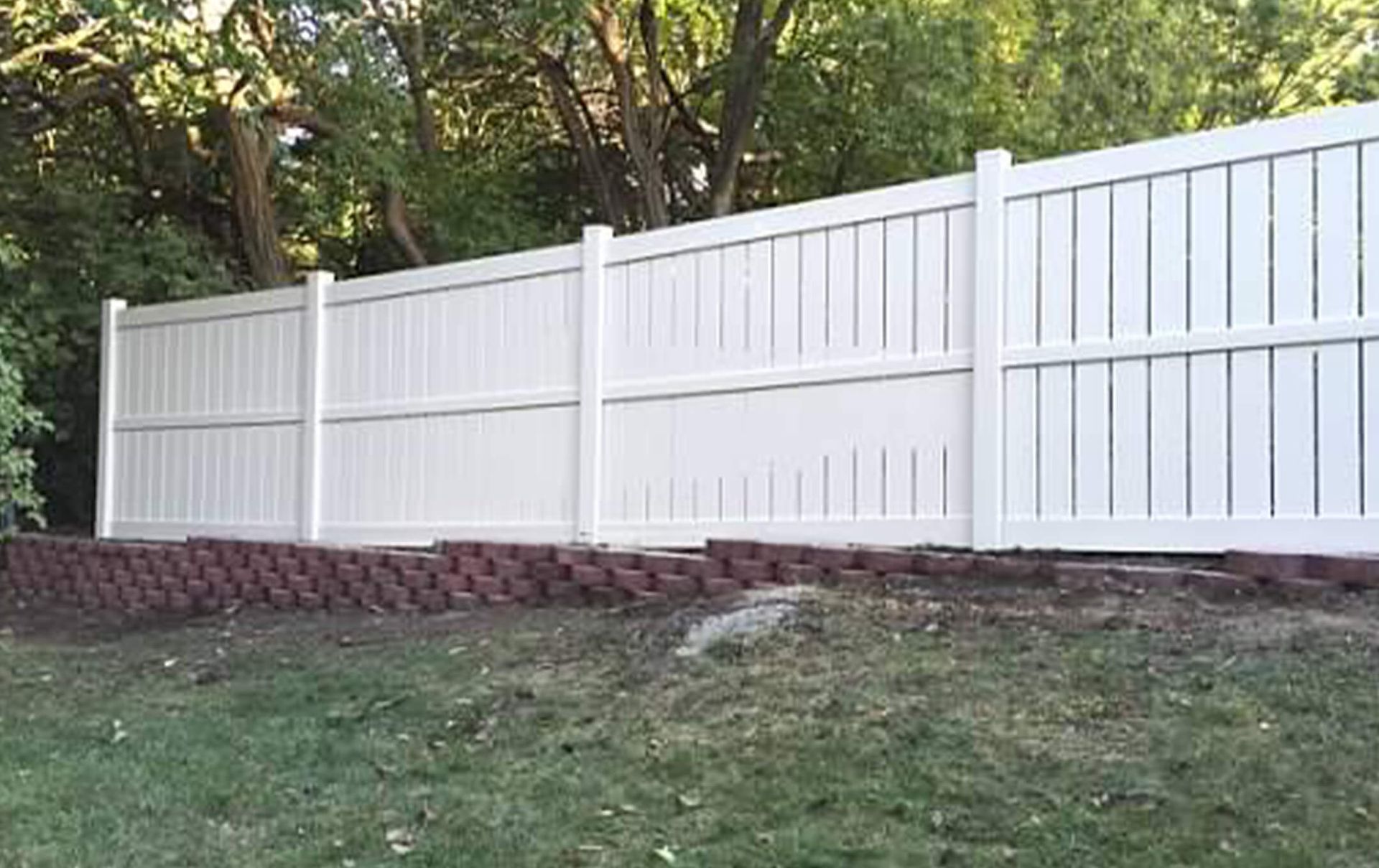 A white vinyl fence surrounds a brick wall in a backyard.