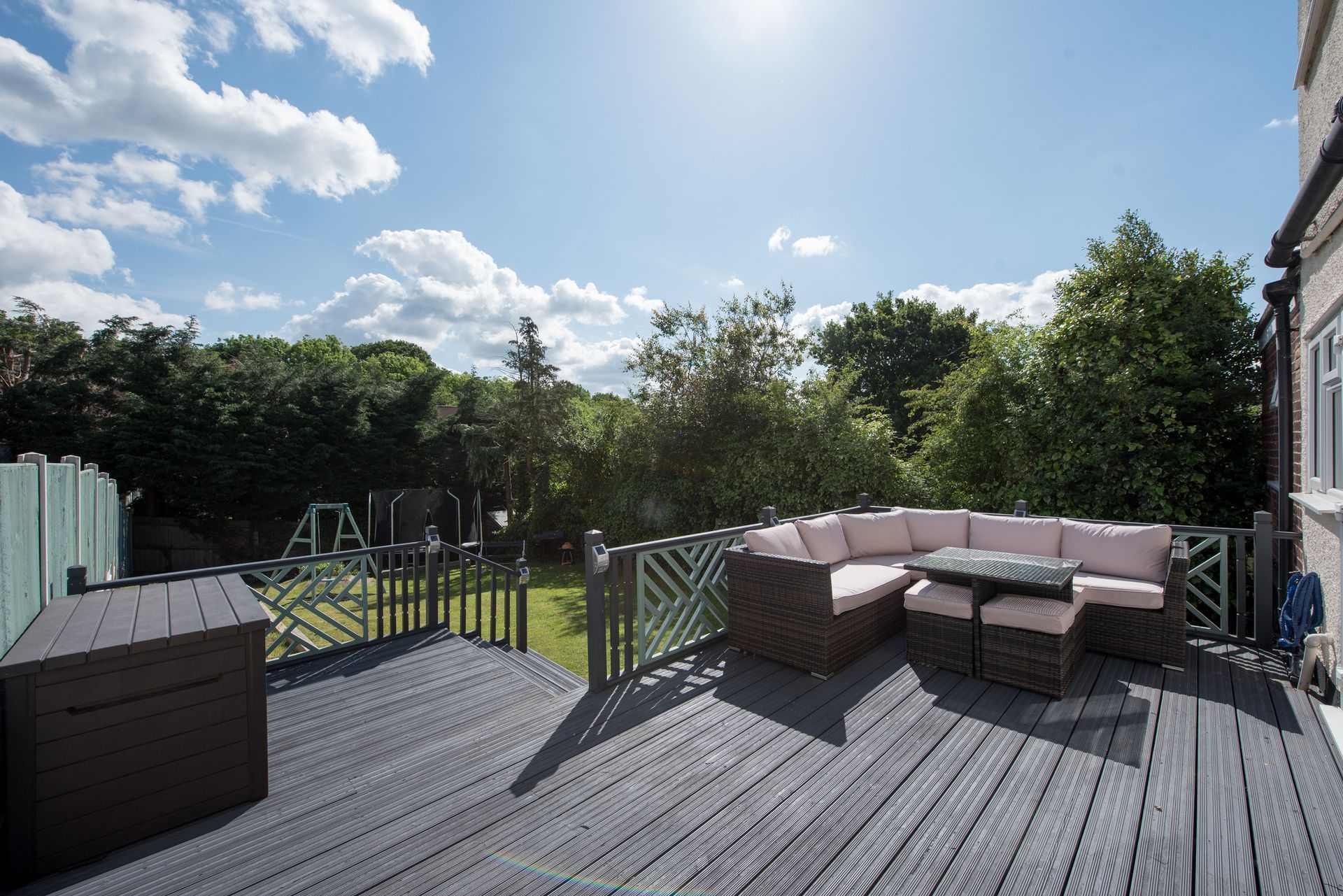 Spacious deck with dark wood flooring, L-shaped sofa, and garden view under a sunny sky.