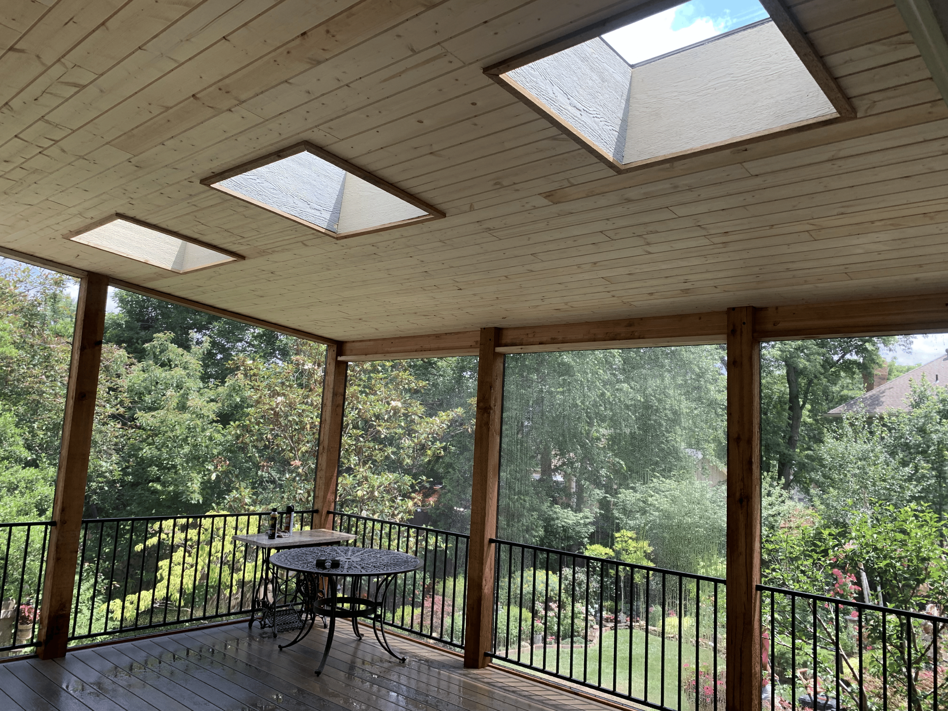 Covered wooden deck with skylights, table and chairs, and a black railing overlooking trees.
