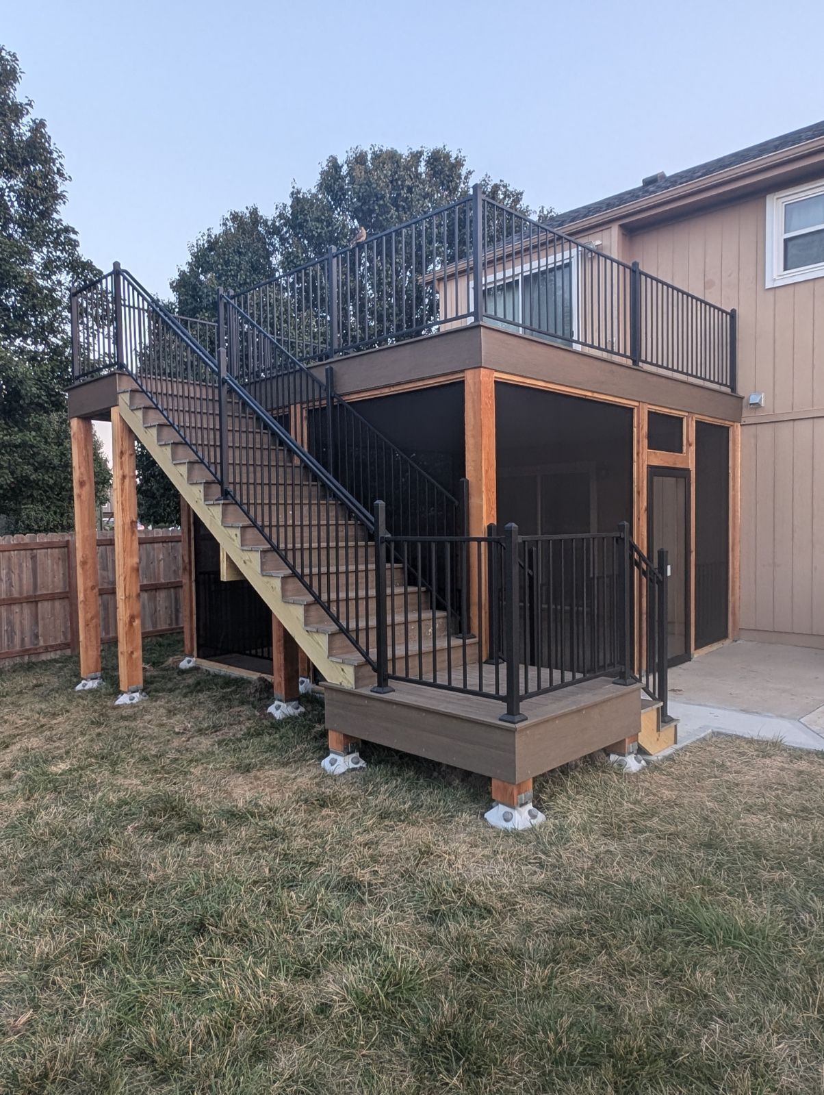 Deck with stairs, black railing, composite decking, and wooden supports. Brown and tan exterior.