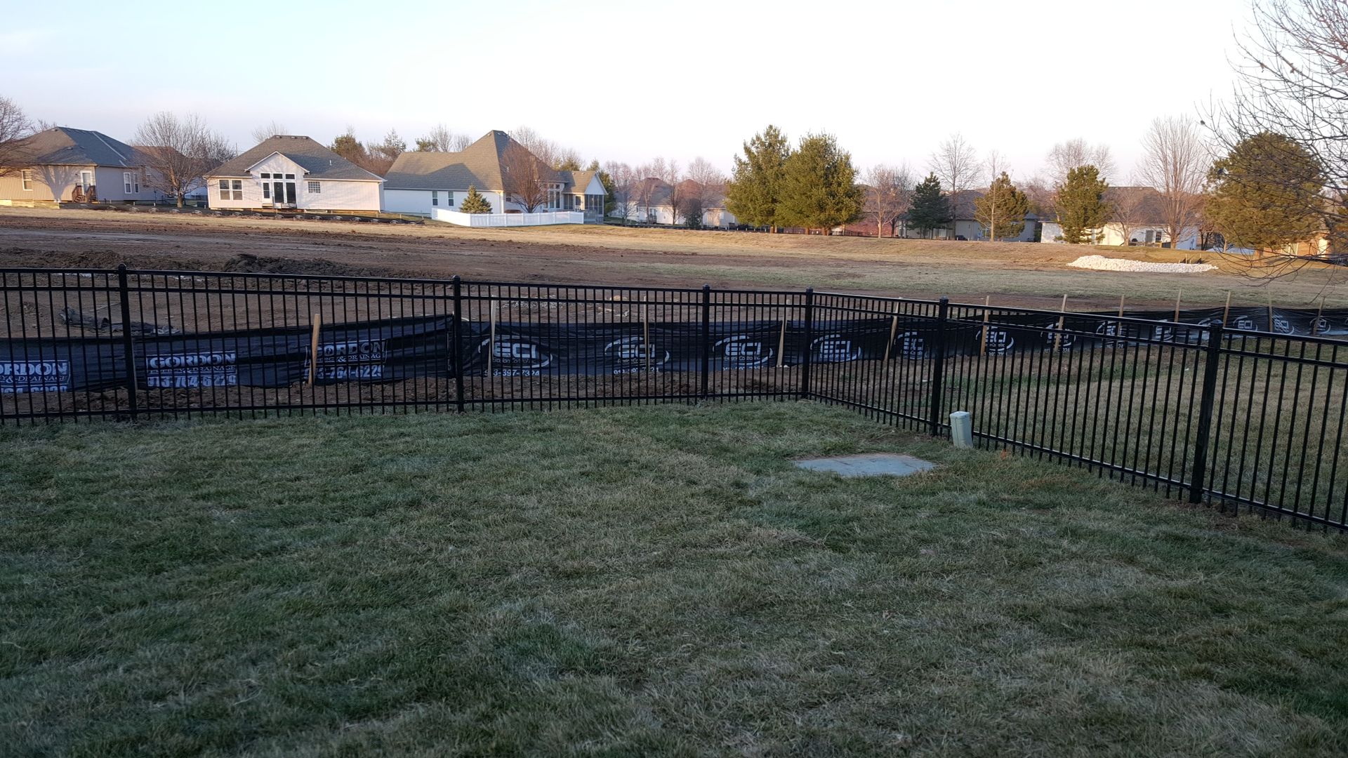 A black fence surrounds a grassy field with houses in the background.
